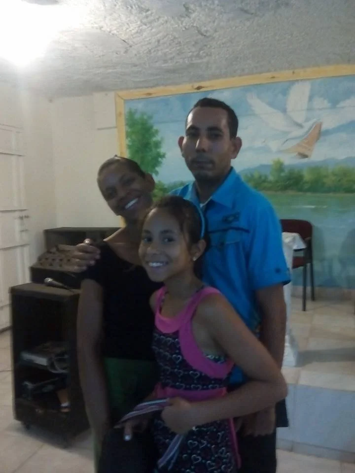 A family of three smiling and posing together indoors, with a mural of a river scene in the background.