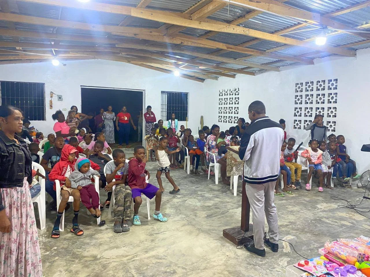 A gathering of children and adults inside a rustic building with a wooden ceiling, metal roof, and white walls. Some children are sitting on white plastic chairs, while others are standing or dancing. An adult is standing at a wooden podium, possibly
