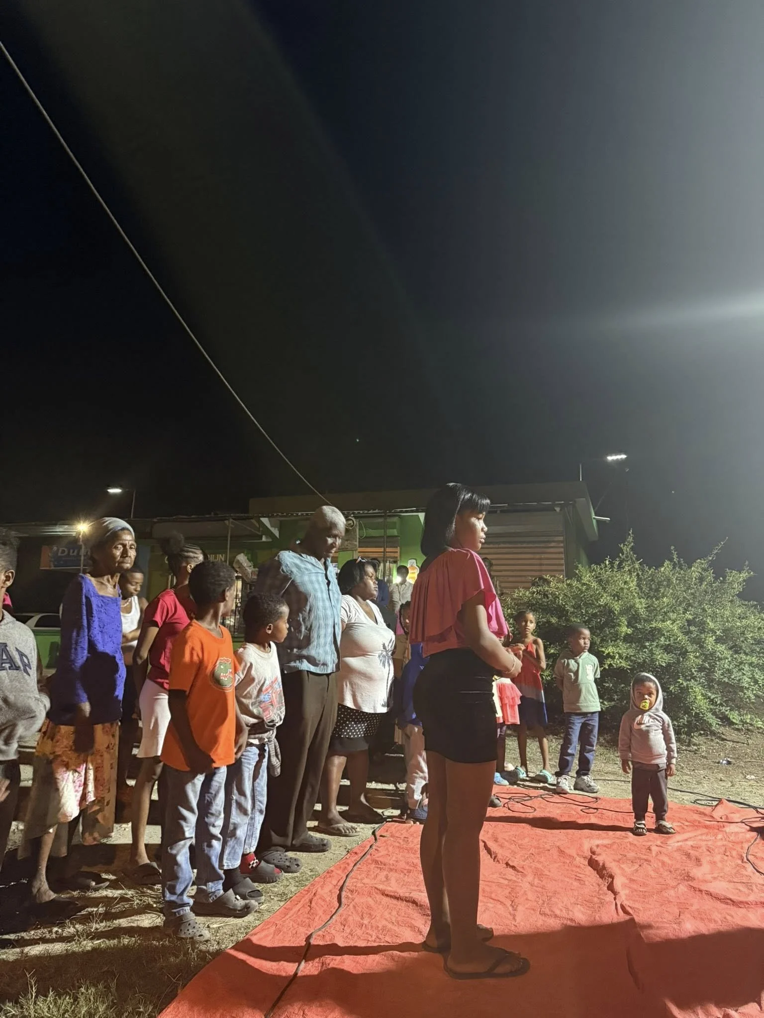 Group of people, including children and adults, gathered outdoors at night on a red carpet, with some looking towards the right. Bright artificial lighting illuminates the scene, with greenery and a small building in the background.