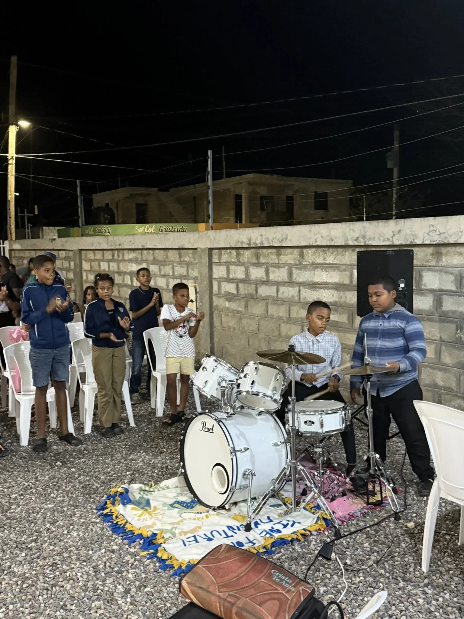 Children and teenagers gathered outdoors at night, some clapping, while two boys play a drum set and a girl plays a cymbal, with a speaker nearby, on a gravel ground enclosed by a brick wall.