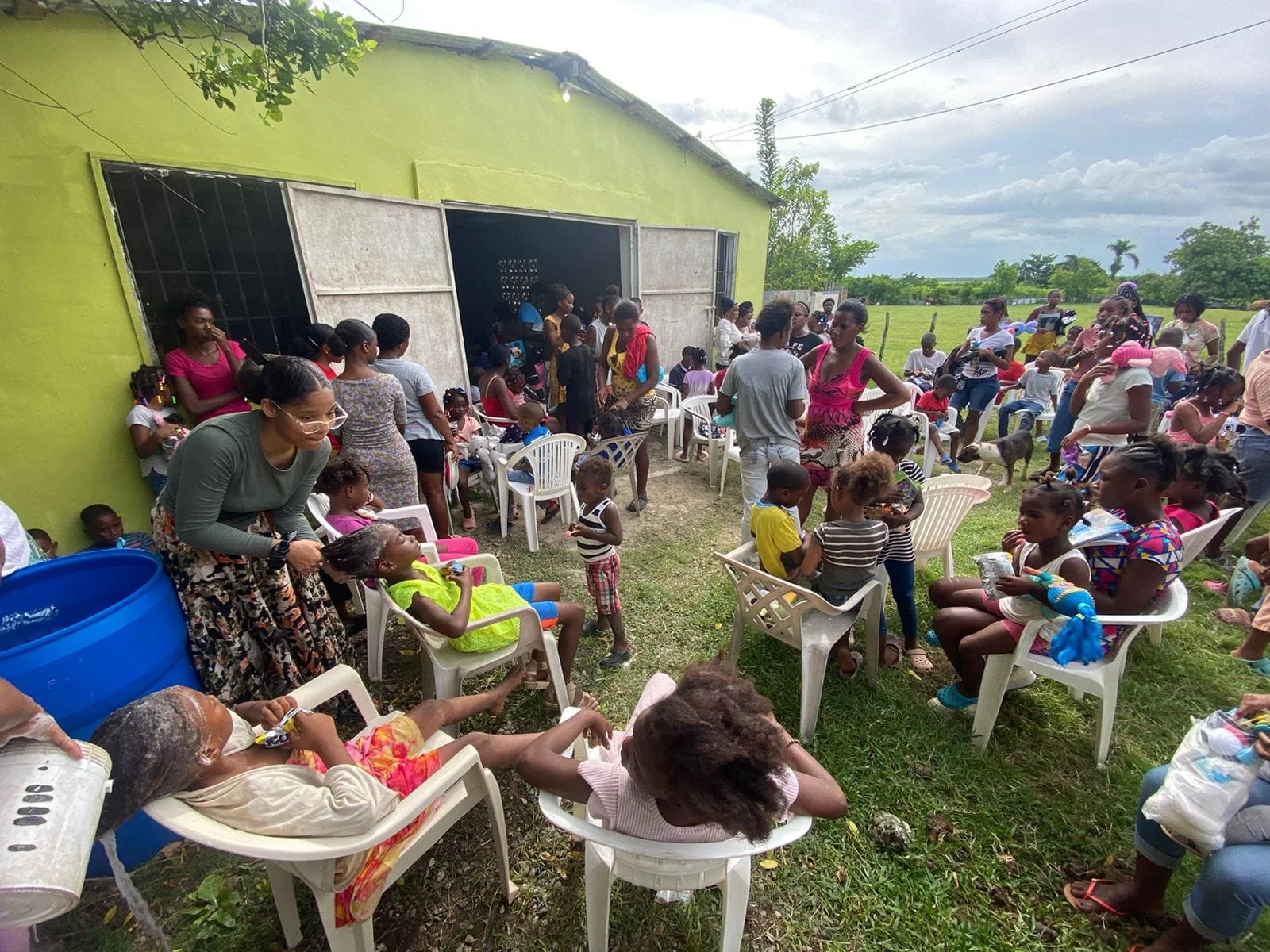 Outdoor gathering of children and women outside a green building, with many seated in white plastic chairs and some standing, in a rural area on a cloudy day.