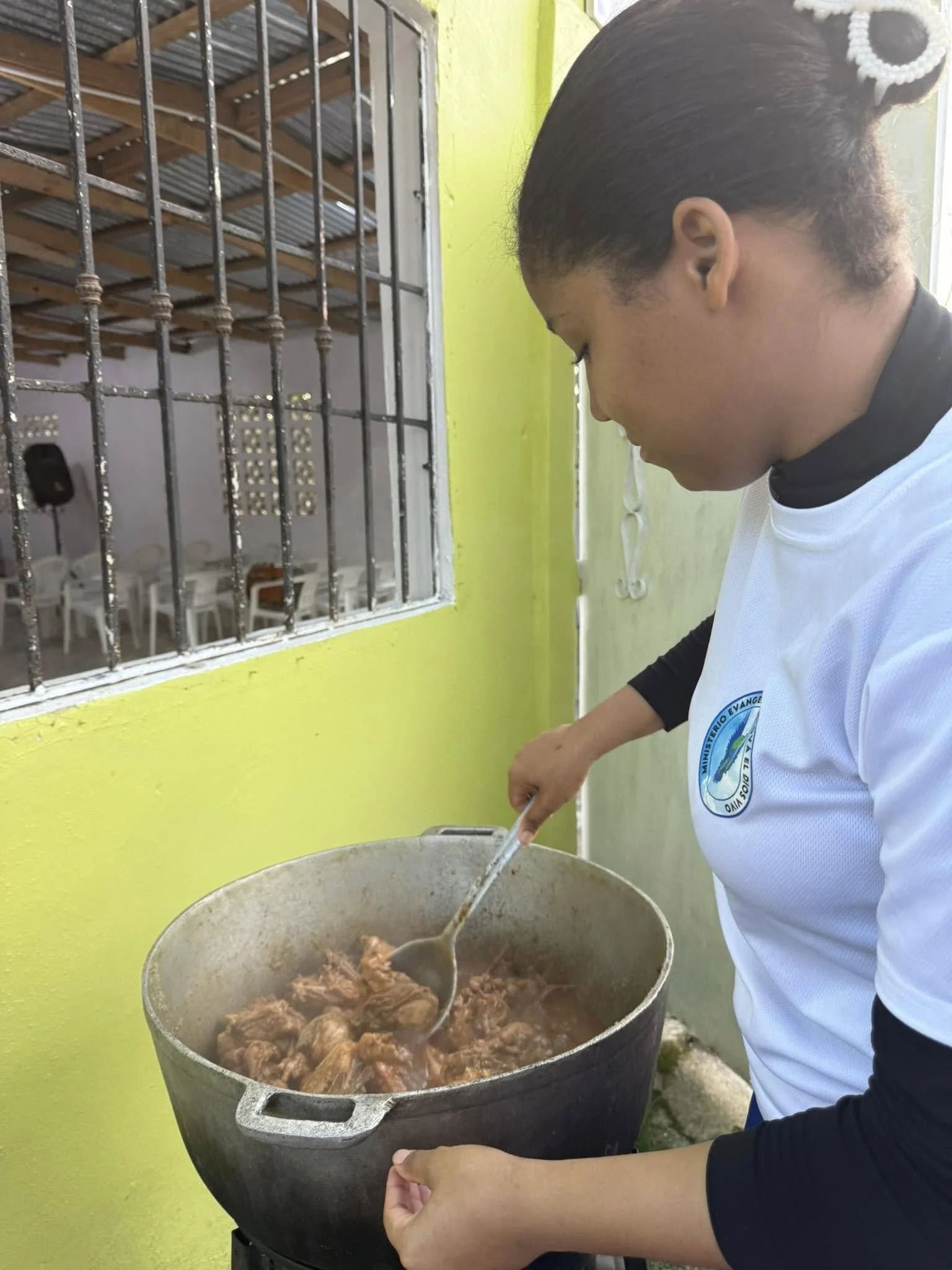 Woman stirring large pot of cooked meat outdoors near yellow wall with barred window.