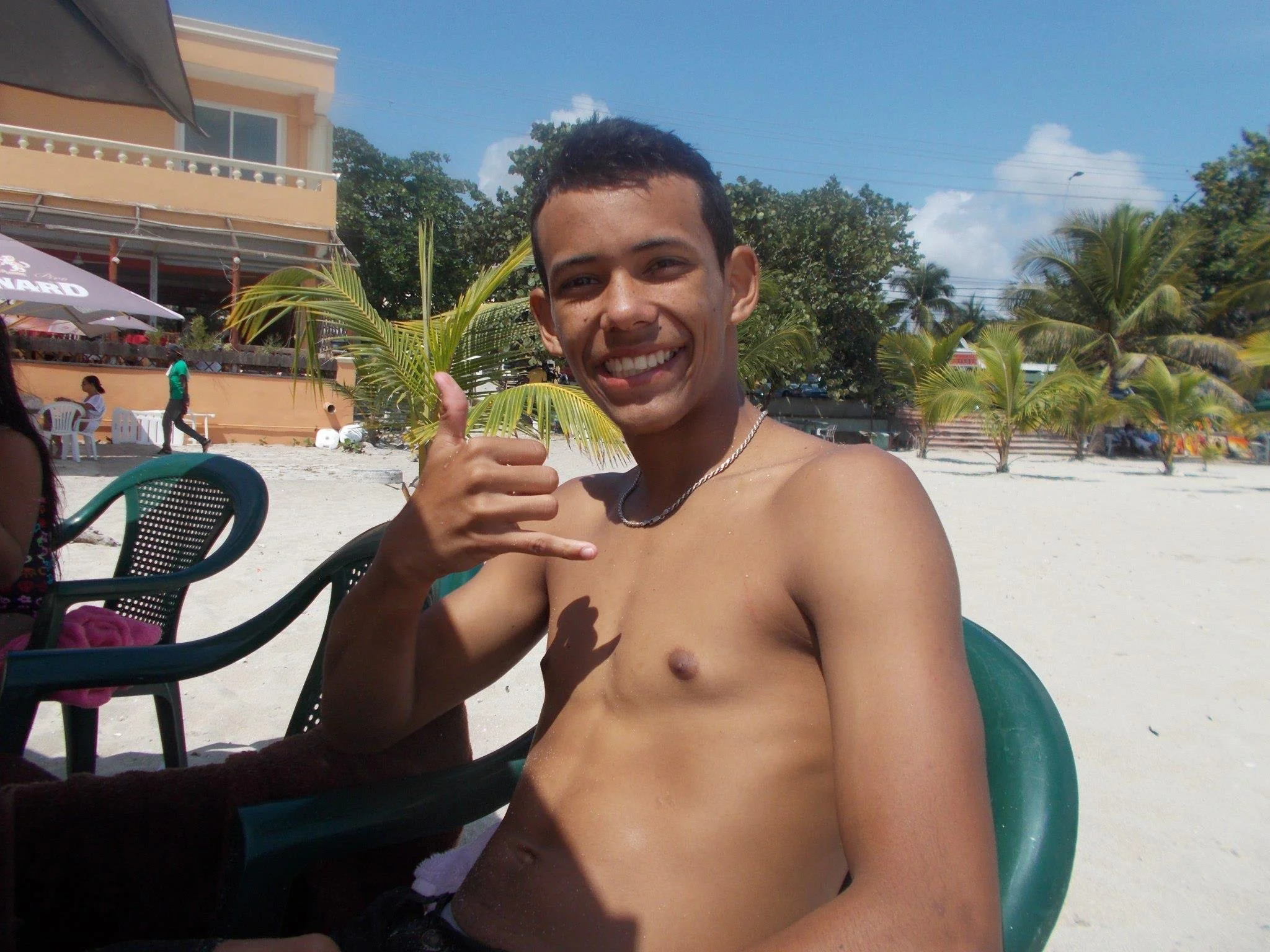A young man smiling and making a phone gesture with his hand on a sunny beach with palm trees, sand, and a building in the background.