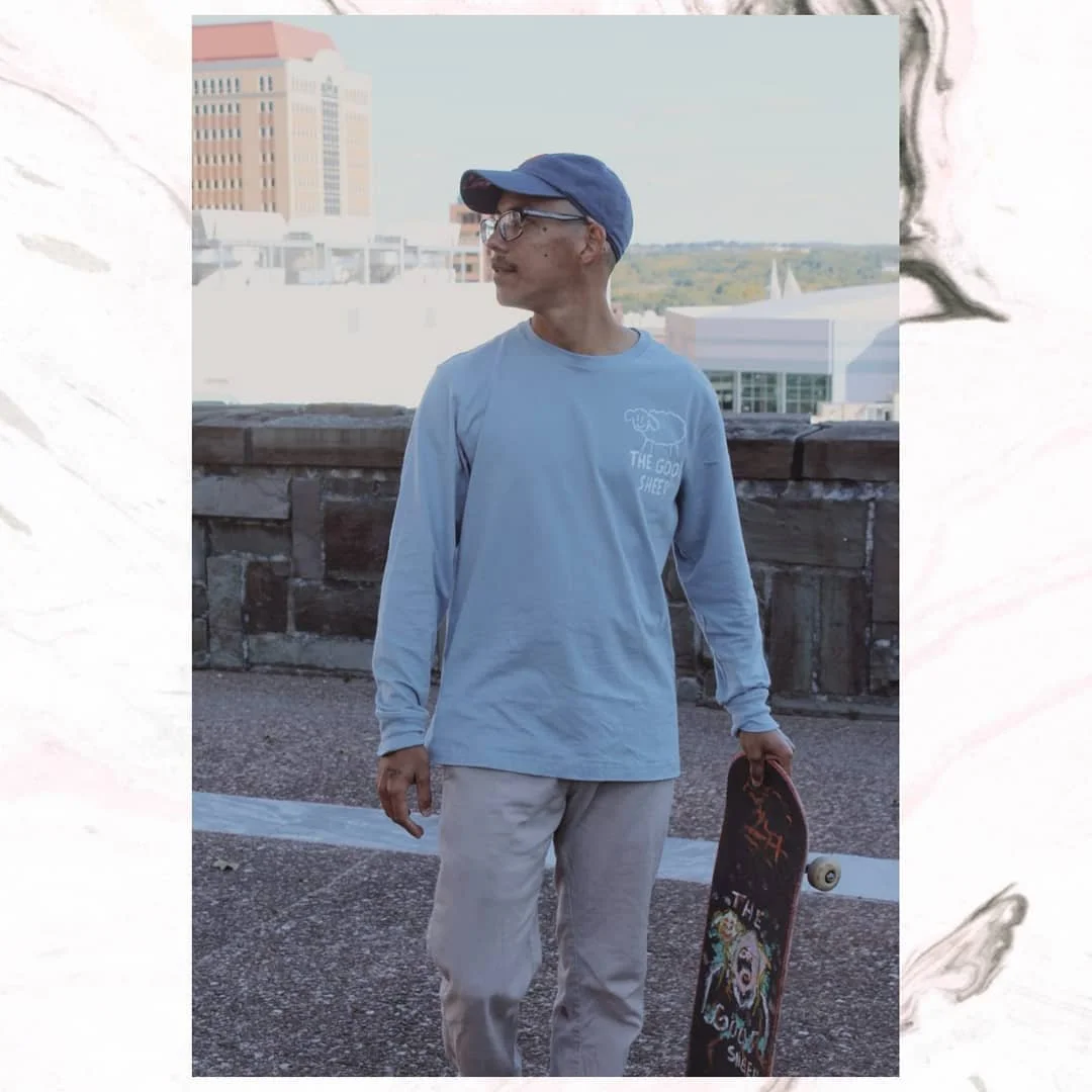 Person wearing glasses, blue cap, and light blue shirt standing outdoors on a rooftop with cityscape and hills in the background, holding a skateboard.