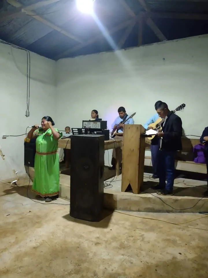 A woman in a bright green dress is singing into a microphone, while four young men play guitar and keyboard on a small stage in a simple room with bare walls and concrete floor.