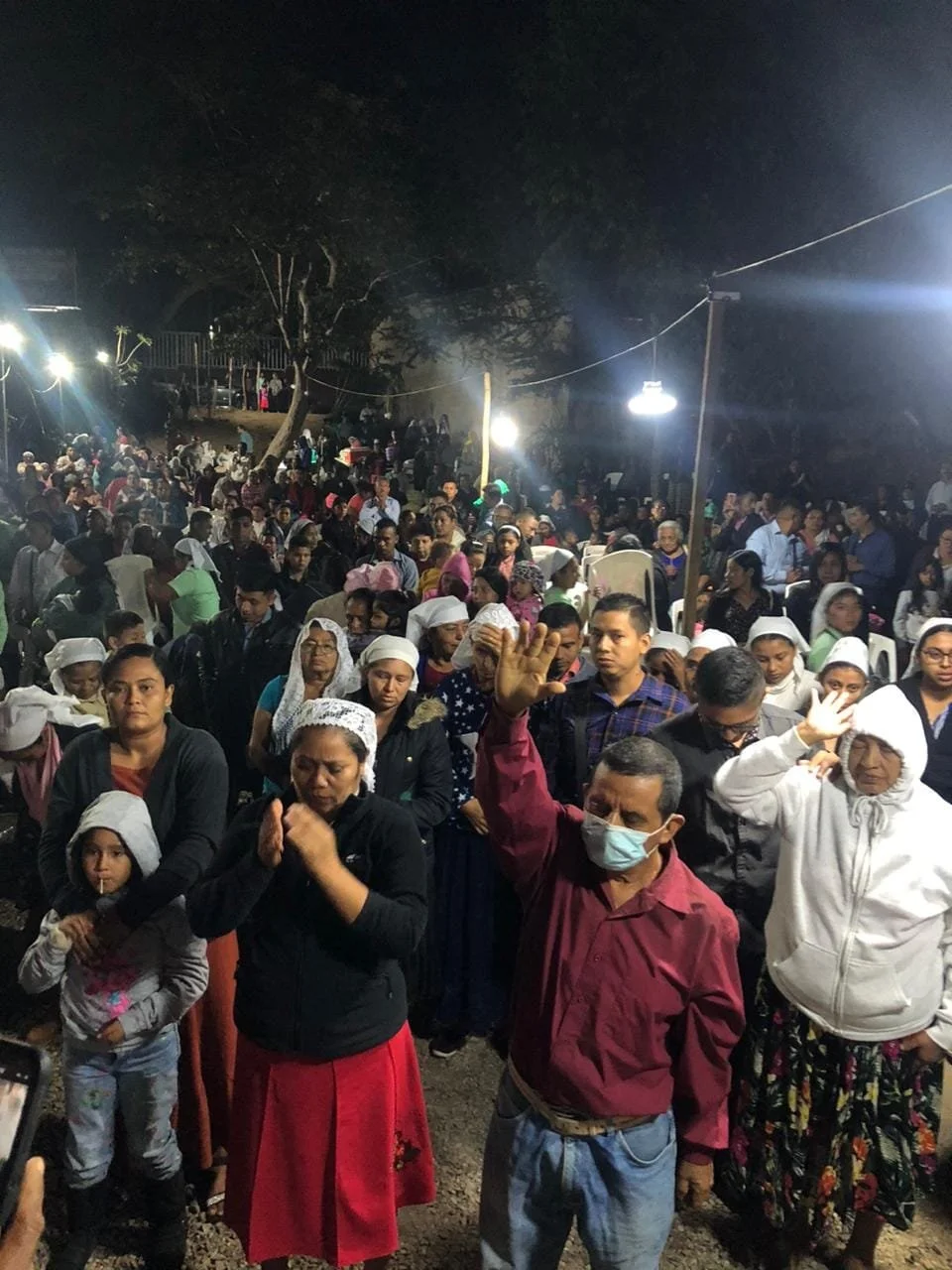 A large crowd gathered outdoors at night, with many people praying and holding their hands up, some wearing head coverings and face masks.