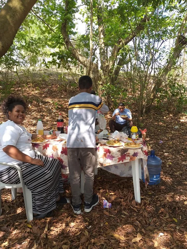 Children enjoying a picnic outdoors in a wooded area with a table set with food, drinks, and snacks surrounded by leafy trees.