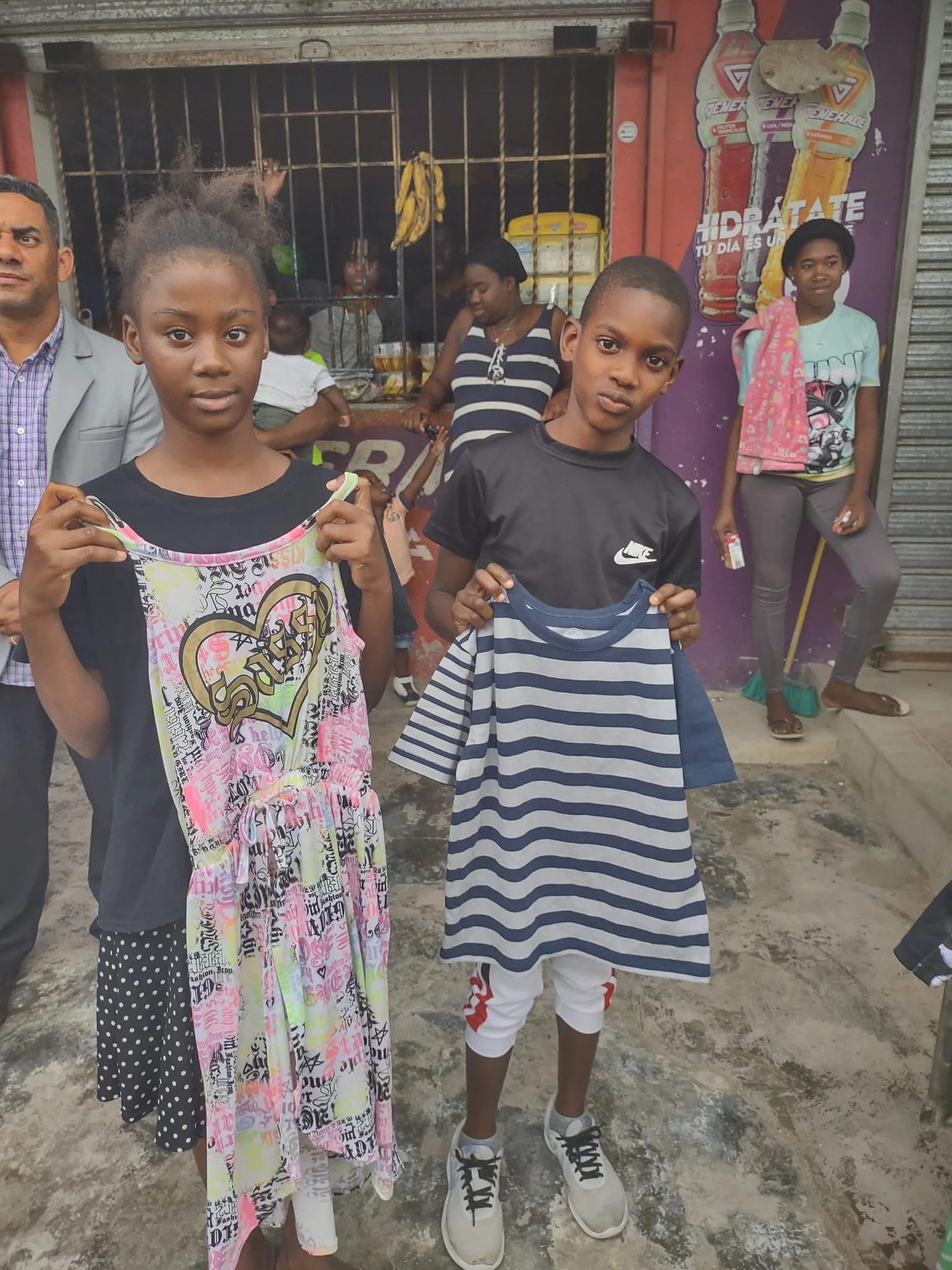Two children standing on a street holding up clothing items, with a small shop and other people in the background.
