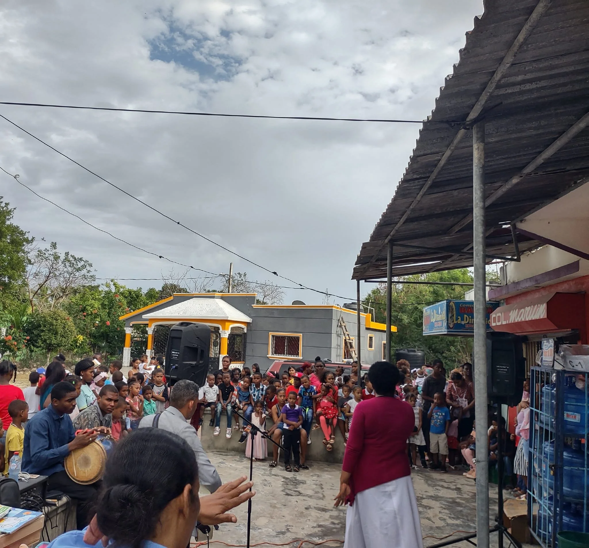 A large group of people, including children, gathered outdoors watching a woman in a red jacket speaking. Musicians are playing instruments nearby, and there are buildings, trees, and cloudy sky in the background.