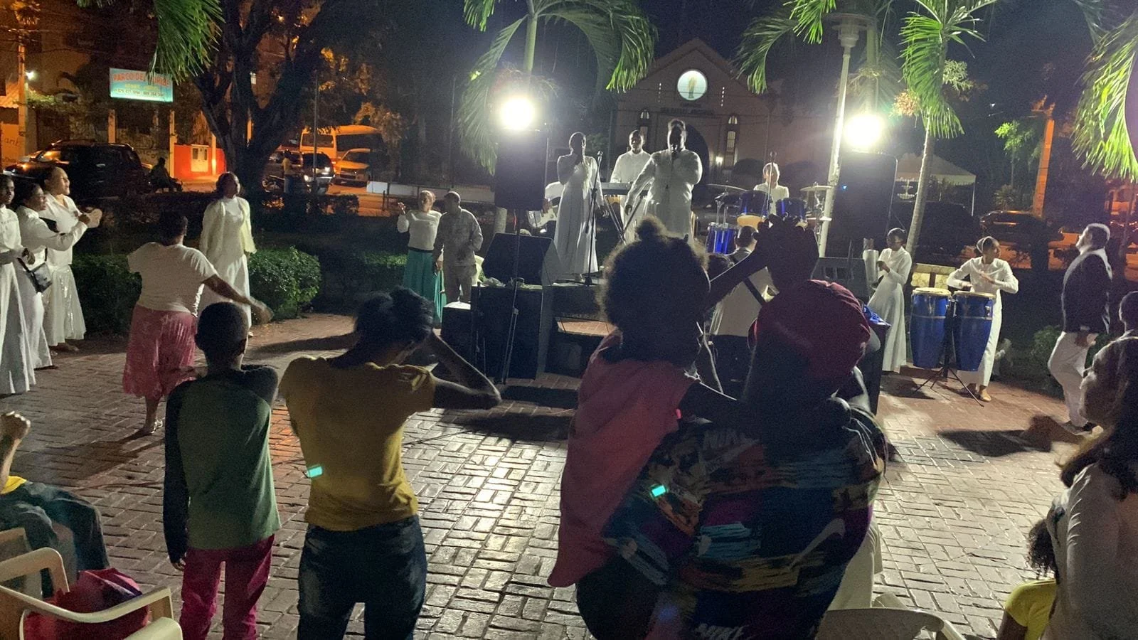 People gathered outdoors at night watching a live outdoor music performance on a small stage, with some dancing and others sitting nearby, illuminated by bright lights.