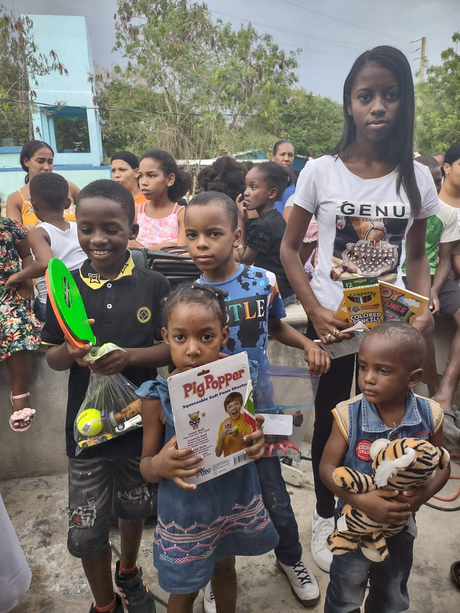 A group of children and adults standing outdoors, some holding toys and books, with trees and a building in the background.