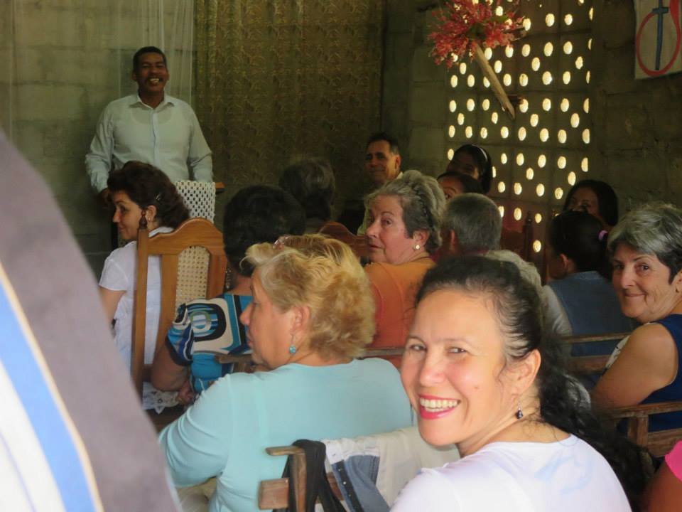 Smiling woman in foreground with black hair pulled back, surrounded by a group of people sitting at a gathering or party indoors.
