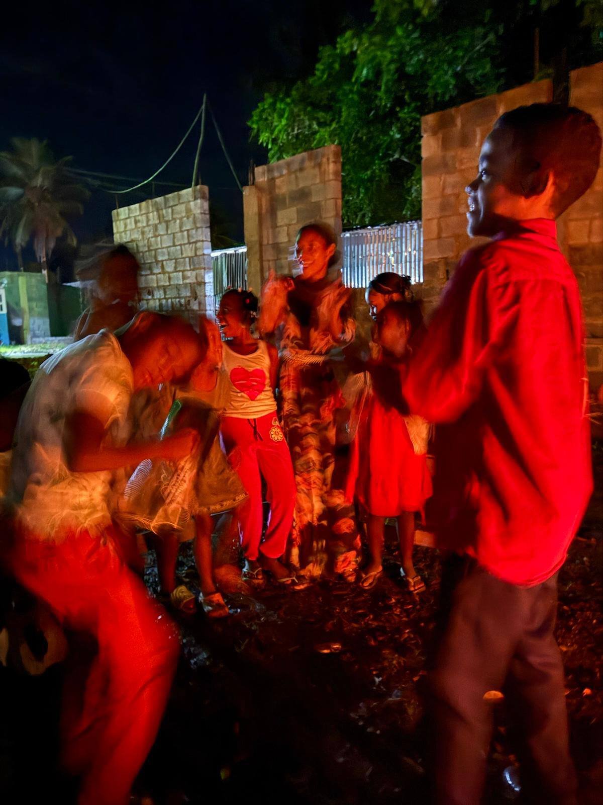 Children and a woman gather outdoors at night, illuminated by red lighting, near a brick wall and trees.