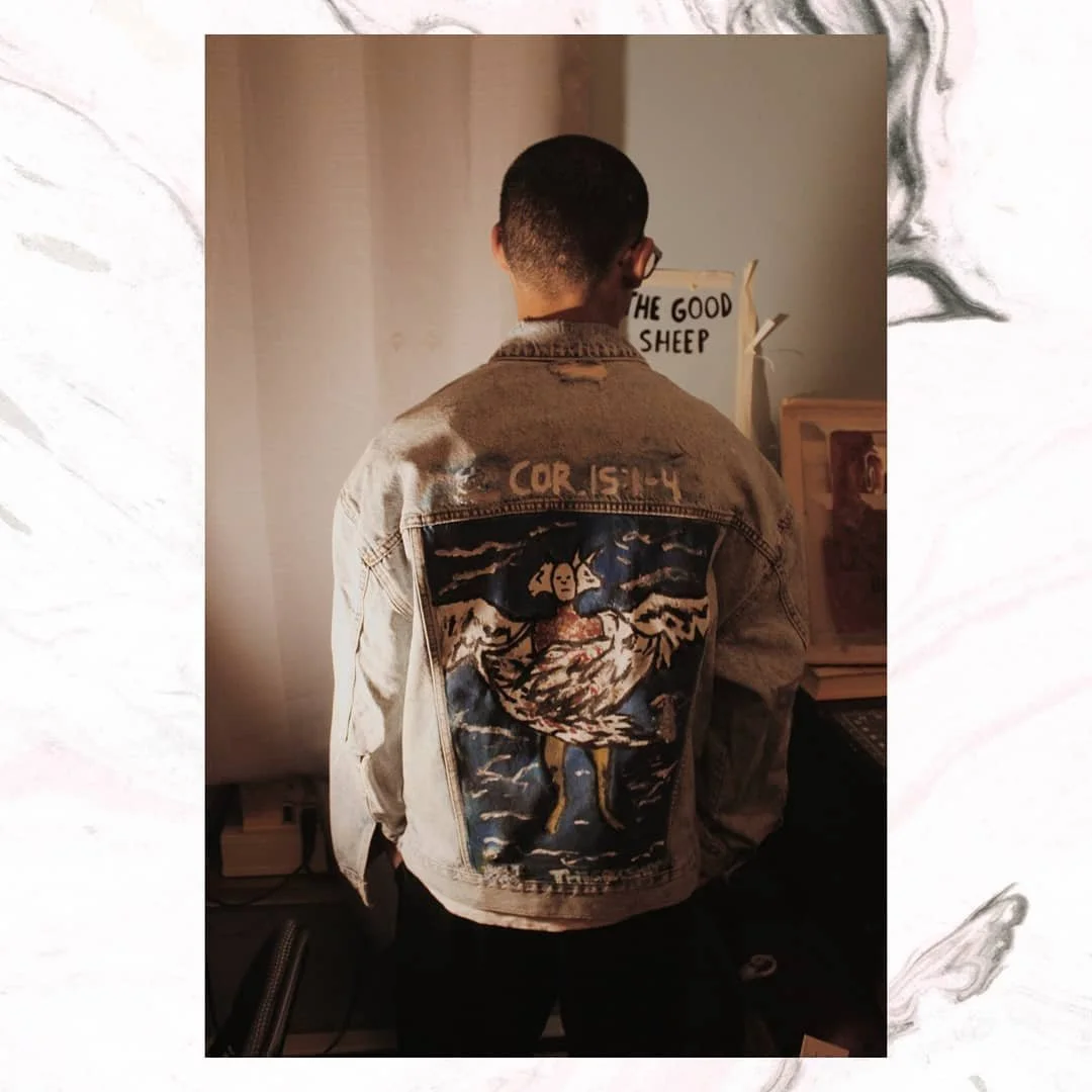 A person with short dark hair wearing glasses and a distressed denim jacket with a large, colorful embroidered patch on the back. The patch features a mythological creature with a woman's face and a lion's body, set against a sea and cloud background