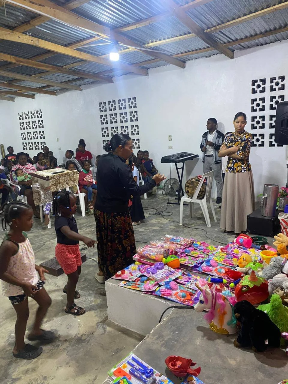 Children and adults gather inside a room for a celebration, with toys and gifts on display in the foreground, and two women speaking at the front.