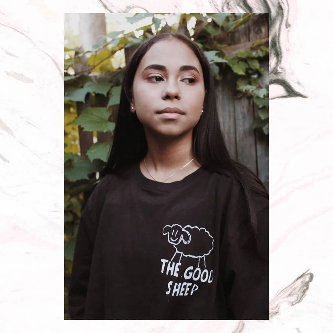 Young woman with long dark hair wearing a black sweatshirt with a sheep graphic and the text "The Good Sheep", standing outdoors with green leaves and a wooden fence in the background.