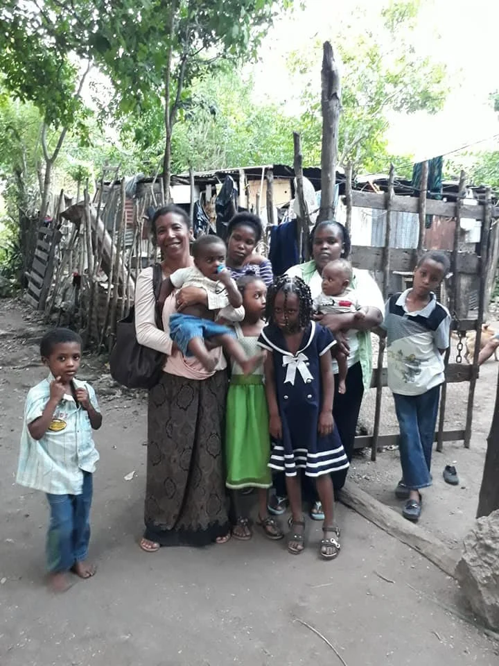 Group of children and two women standing outdoors in front of a wooden fence and a makeshift shelter, surrounded by trees.
