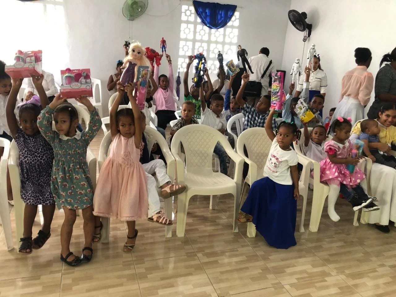 Children sitting and standing in a room, holding toys and gifts, some with raised hands, during a celebration or party.
