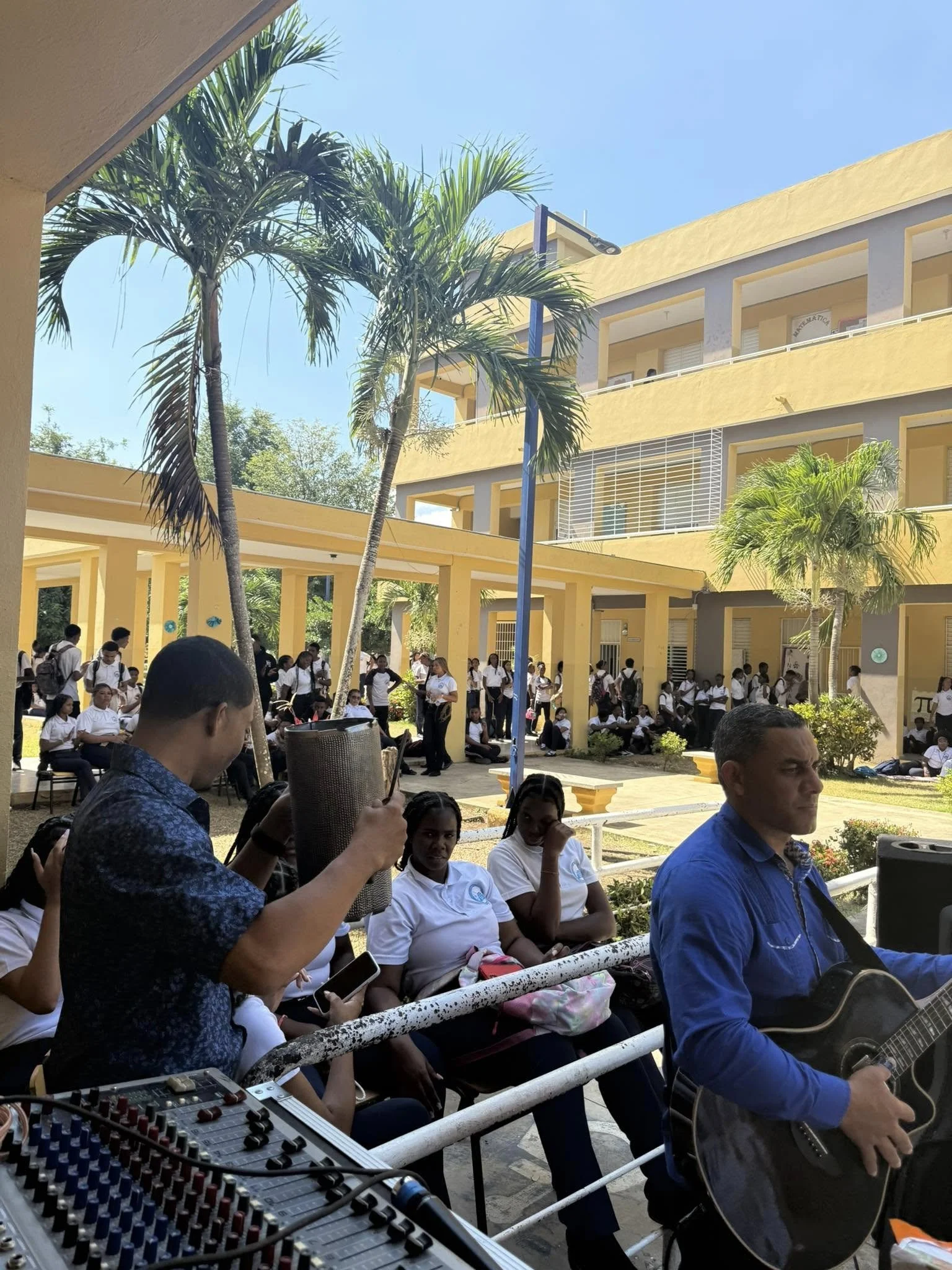 Students gathered outside a yellow school building, some sitting and some standing, with a man playing guitar and another man adjusting sound equipment. Palm trees and clear blue sky are visible.