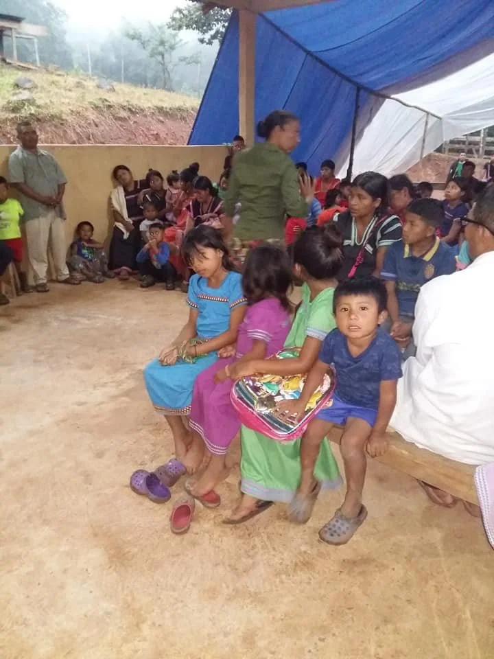 A group of children and adults sitting and standing in an outdoor setting with a makeshift blue and white tent overhead.