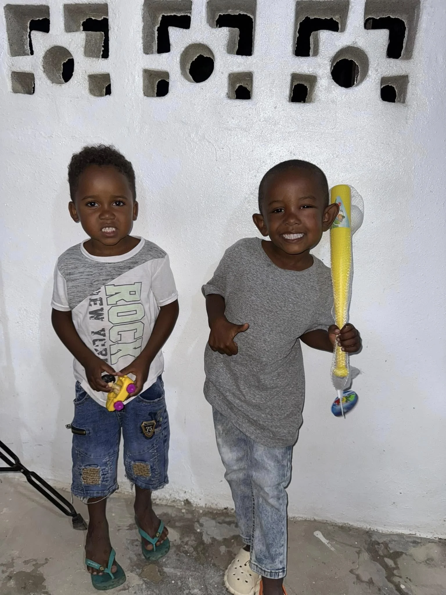 Two young boys standing against a white wall with geometric and circular cutouts, smiling at the camera, holding toys, one with a small yellow toy car and the other with a bubble wand.