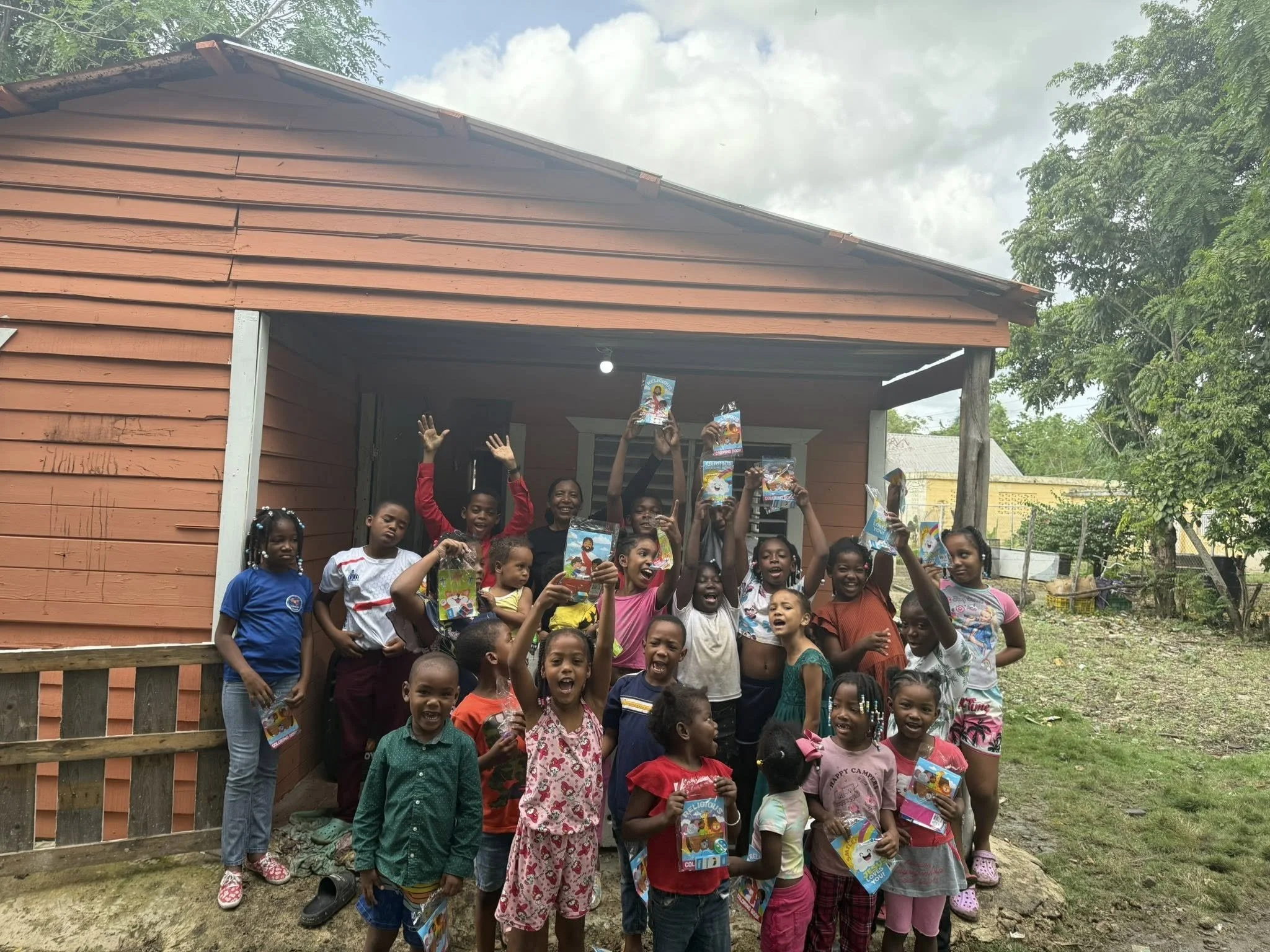 A group of children and a woman standing in front of a cottage, holding up colorful packages, and smiling outdoors.
