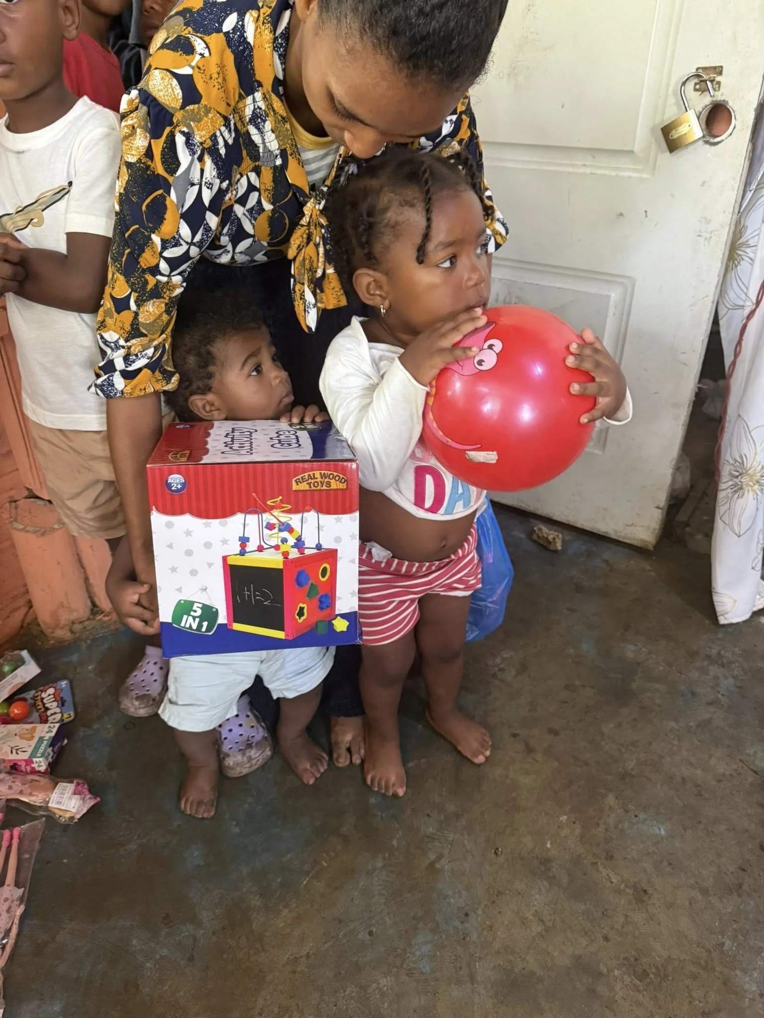 A woman is holding a young girl with braided hair, who is holding a red balloon. Another child is behind them, partially obscured, holding a toy or decoration. A child on the left is holding a toy car. The scene appears to be in a modest indoor setti