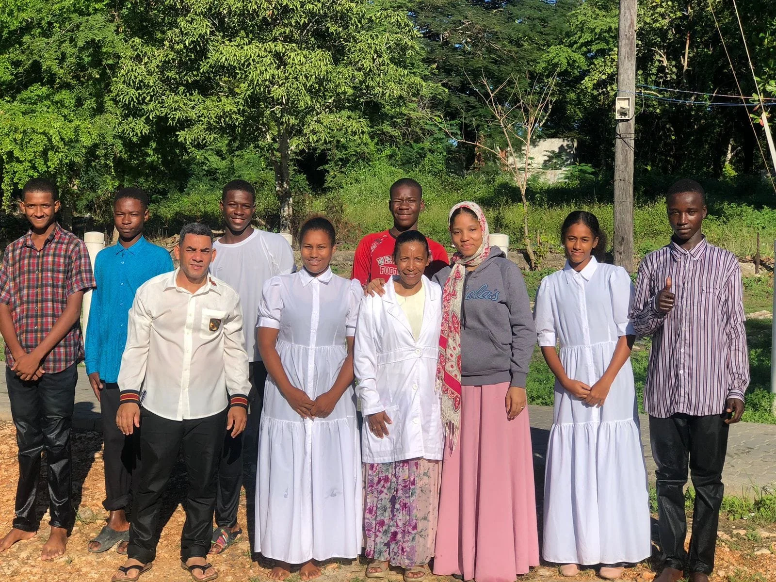 A group of ten people, including women and men, standing outdoors on a sunny day with green trees and power lines in the background. Some of the women are wearing white dresses, and some men are in casual shirts. Everyone is smiling, and one person o