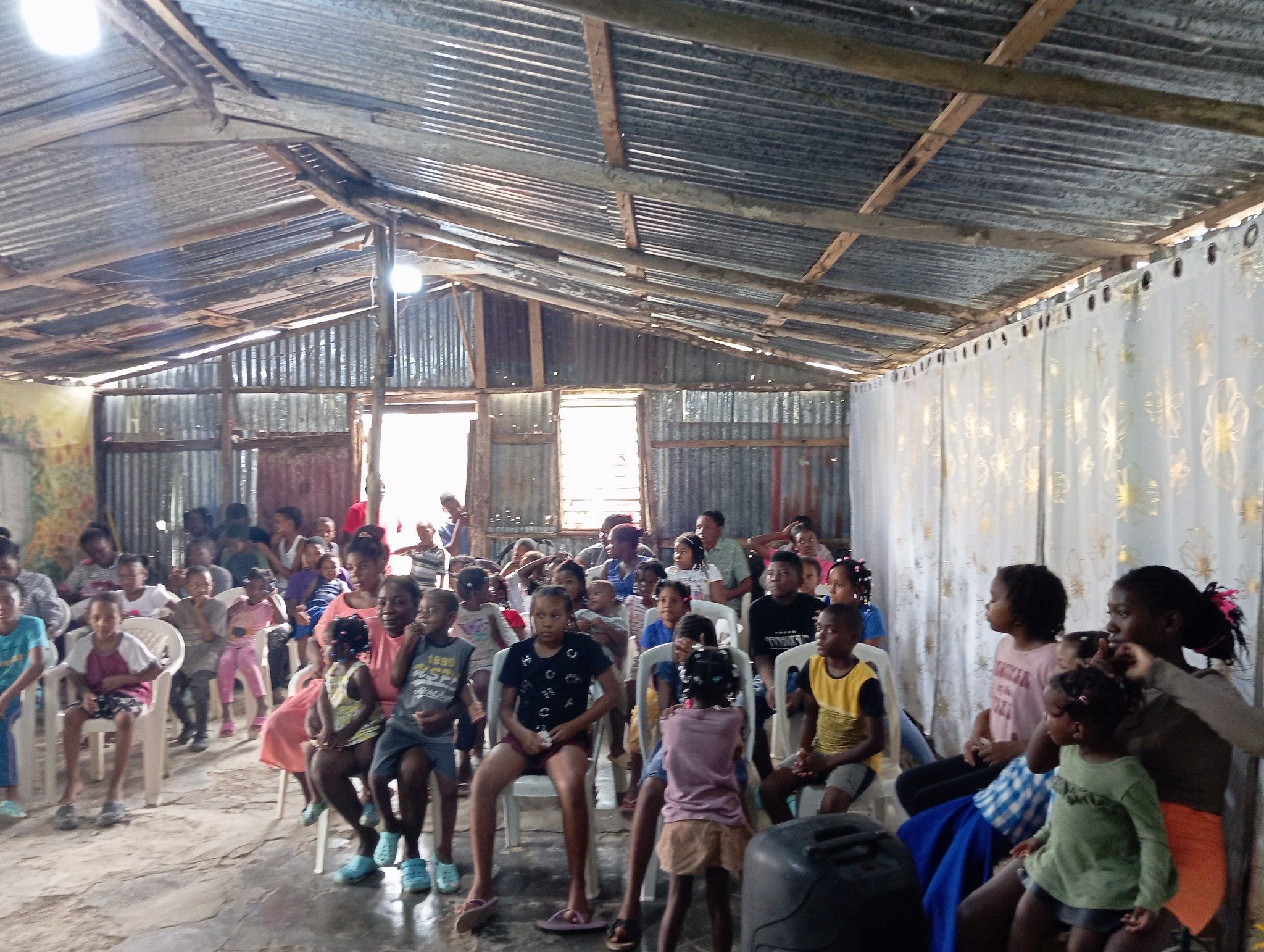 A group of children and adults seated inside a rustic, open wooden and corrugated metal structure, with some sitting on plastic chairs and others standing, engaged in an activity or event.
