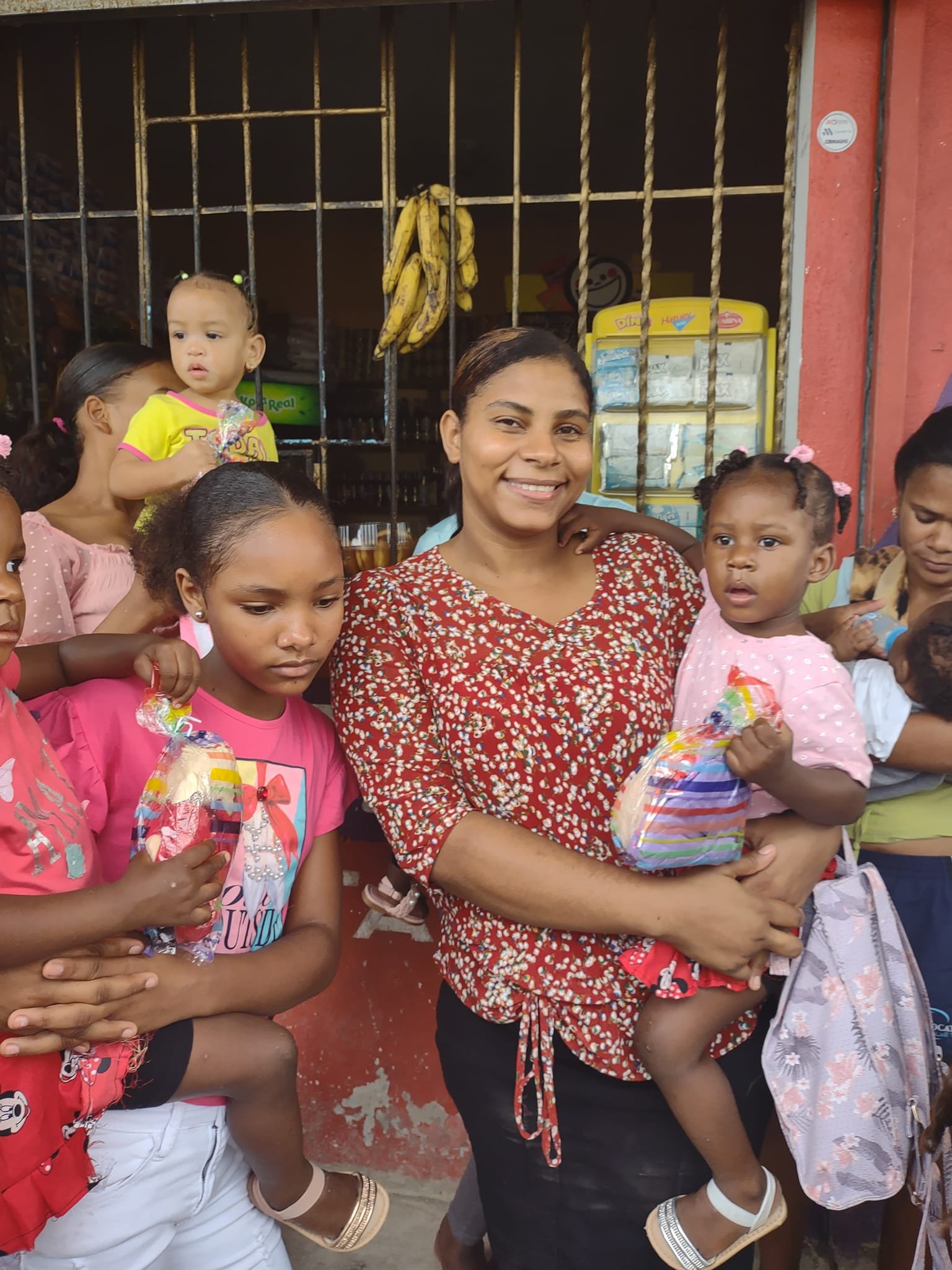 A group of women and children standing outside a shop for a celebration, with one woman holding a young girl. Some children are holding small gift bags.