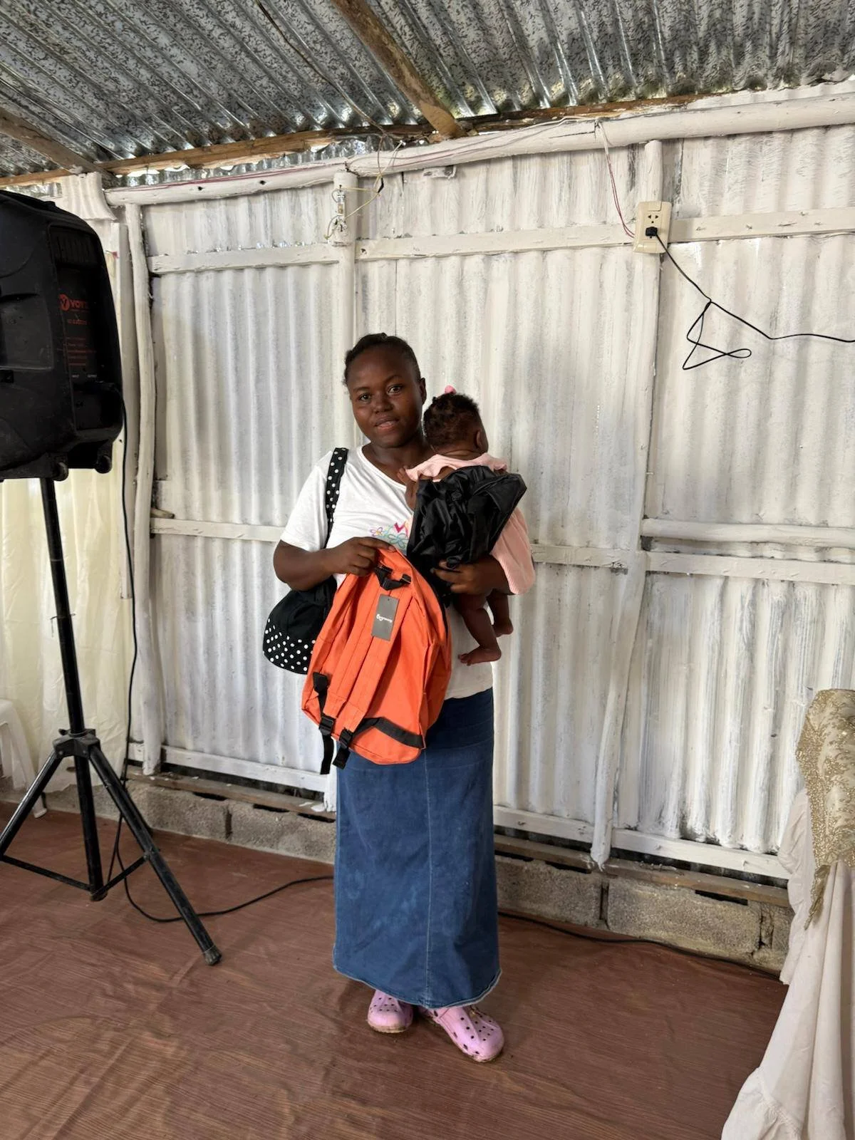 A woman holding a small child stands indoors in front of a corrugated metal wall. She is holding an orange backpack and wearing pink Crocs and a denim skirt.