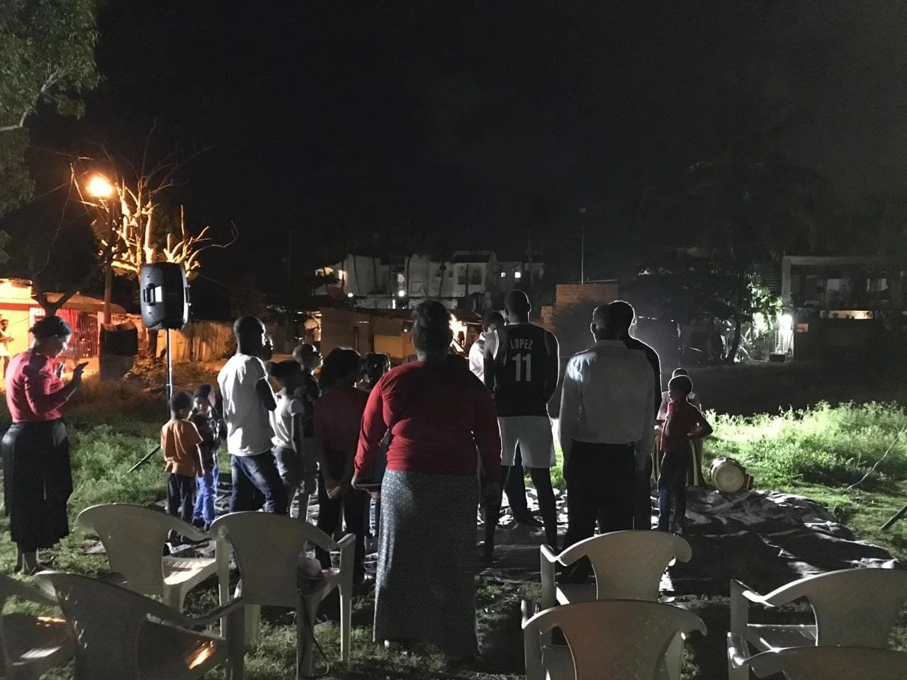 A group of people, including children and adults, gather outdoors at night around a small area with a tarp, illuminated by a nearby streetlight and some additional lighting. Some chairs are visible in the foreground.