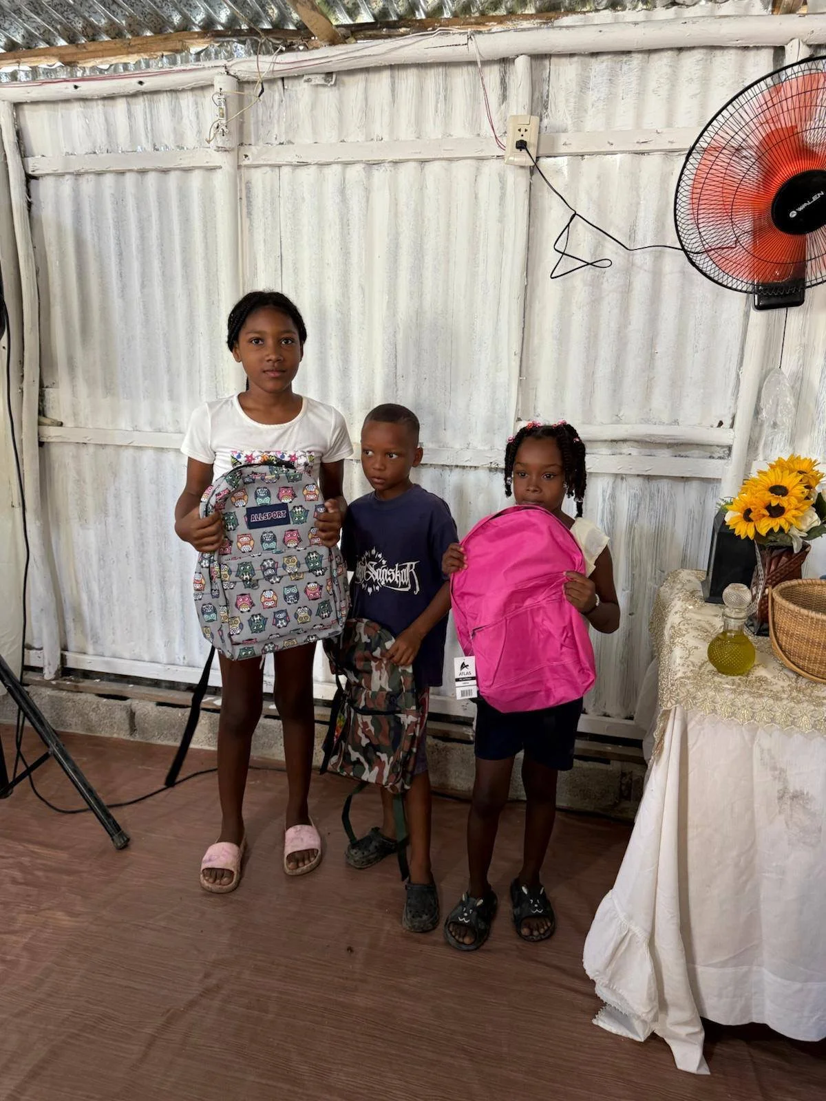 Three children standing indoors holding backpacks, with a white corrugated metal wall in the background and a table with a sunflower arrangement on the right.