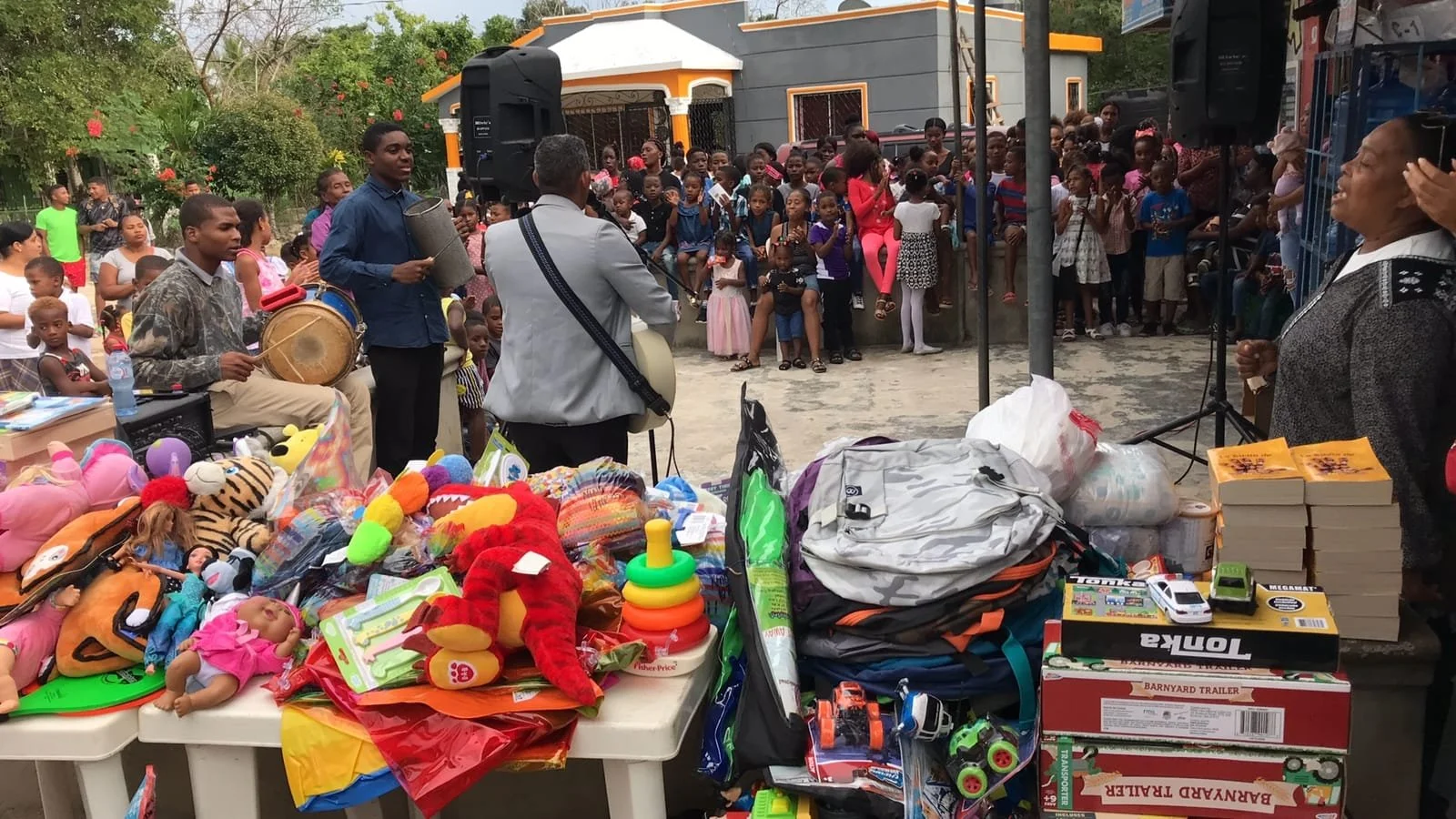 A community event with a large gathering of children and adults. There are tables with toys, books, and other items for sale or display in the foreground. A group of musicians playing drums and guitars are performing in front of a crowd of children a