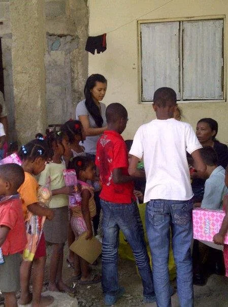 Children and women gathered outside a house with a woman handing out items at a table