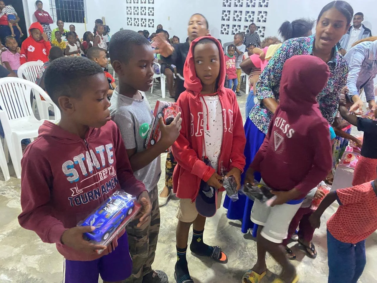 Children and adults gathered in an indoor space, some children holding toys and gifts, with chairs and a white wall in the background, suggesting a celebration or party.