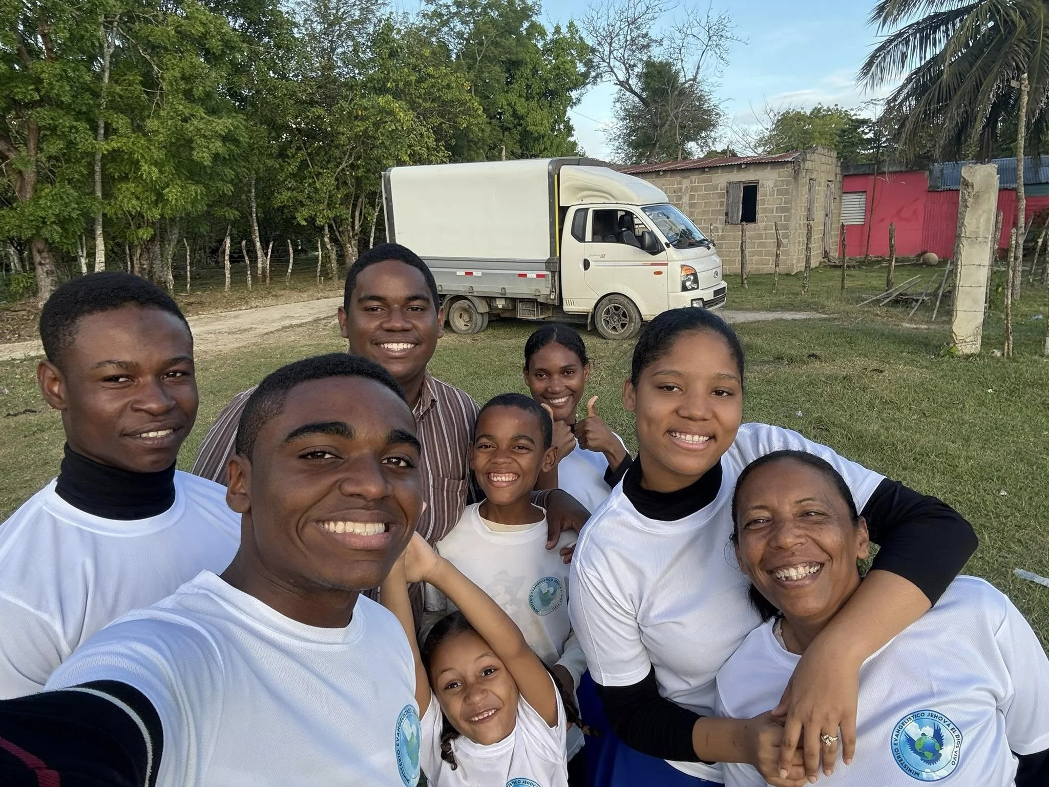 Group of smiling children and adults taking a selfie outdoors with trees, a white truck, and small buildings in the background.