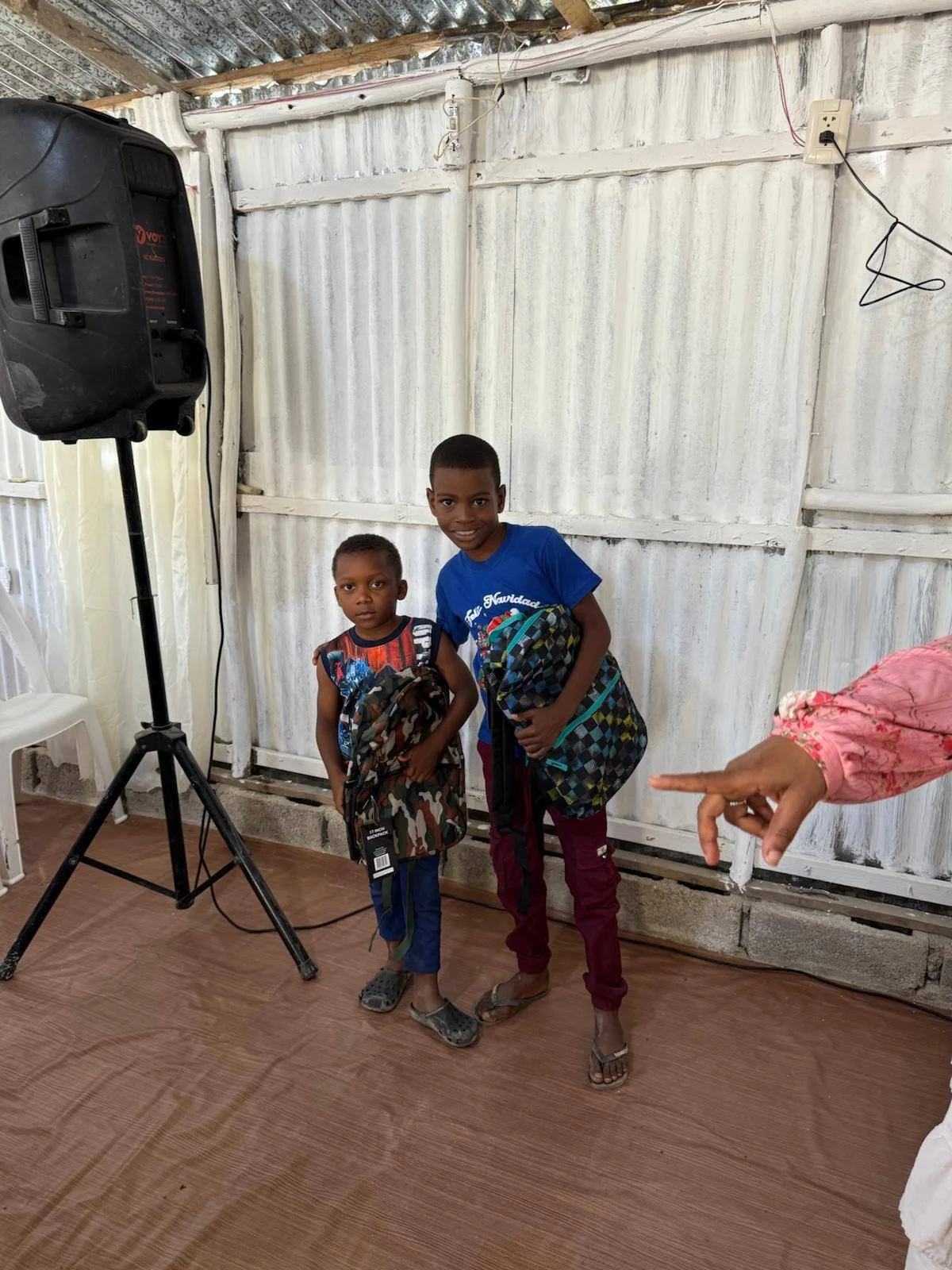 Two young boys standing indoors, both holding backpacks. The boy on the left is smaller, wearing a sleeveless shirt and blue pants, while the taller boy on the right is wearing a blue T-shirt and maroon pants. They are posing for the photo, with a wh