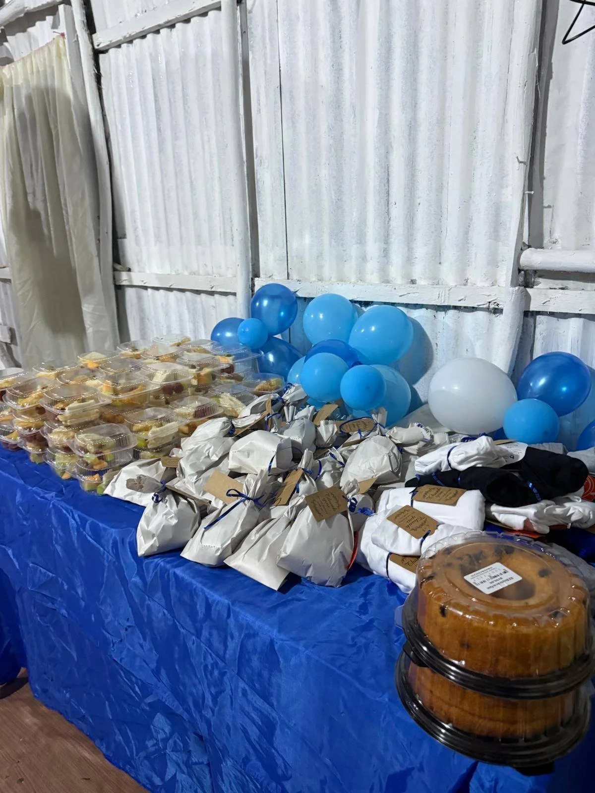 Party table decorated with blue and white balloons, with various wrapped gift bags, plastic containers of cake, and plate of cookies.
