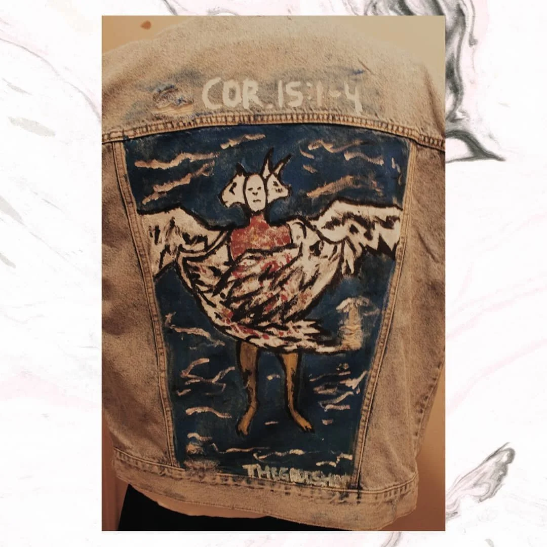 Back of a denim jacket with a painted chicken, clouds, and mountains, with the text 'COR 15-14' and 'THANKS' written on it.