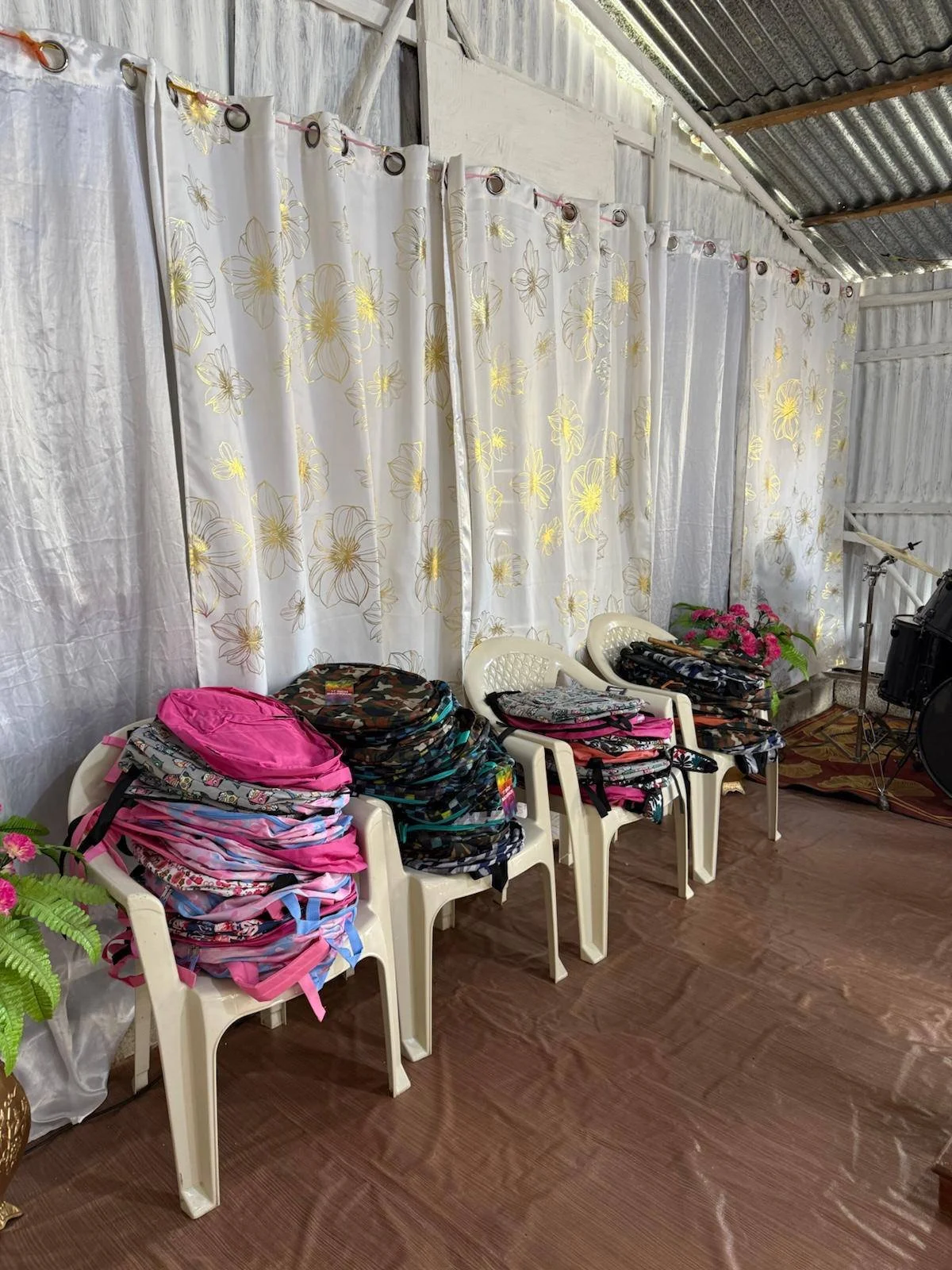 Four plastic chairs lined up against a white curtain backdrop with backpacks and bags stacked on each chair, with potted plants and a musical instrument nearby.