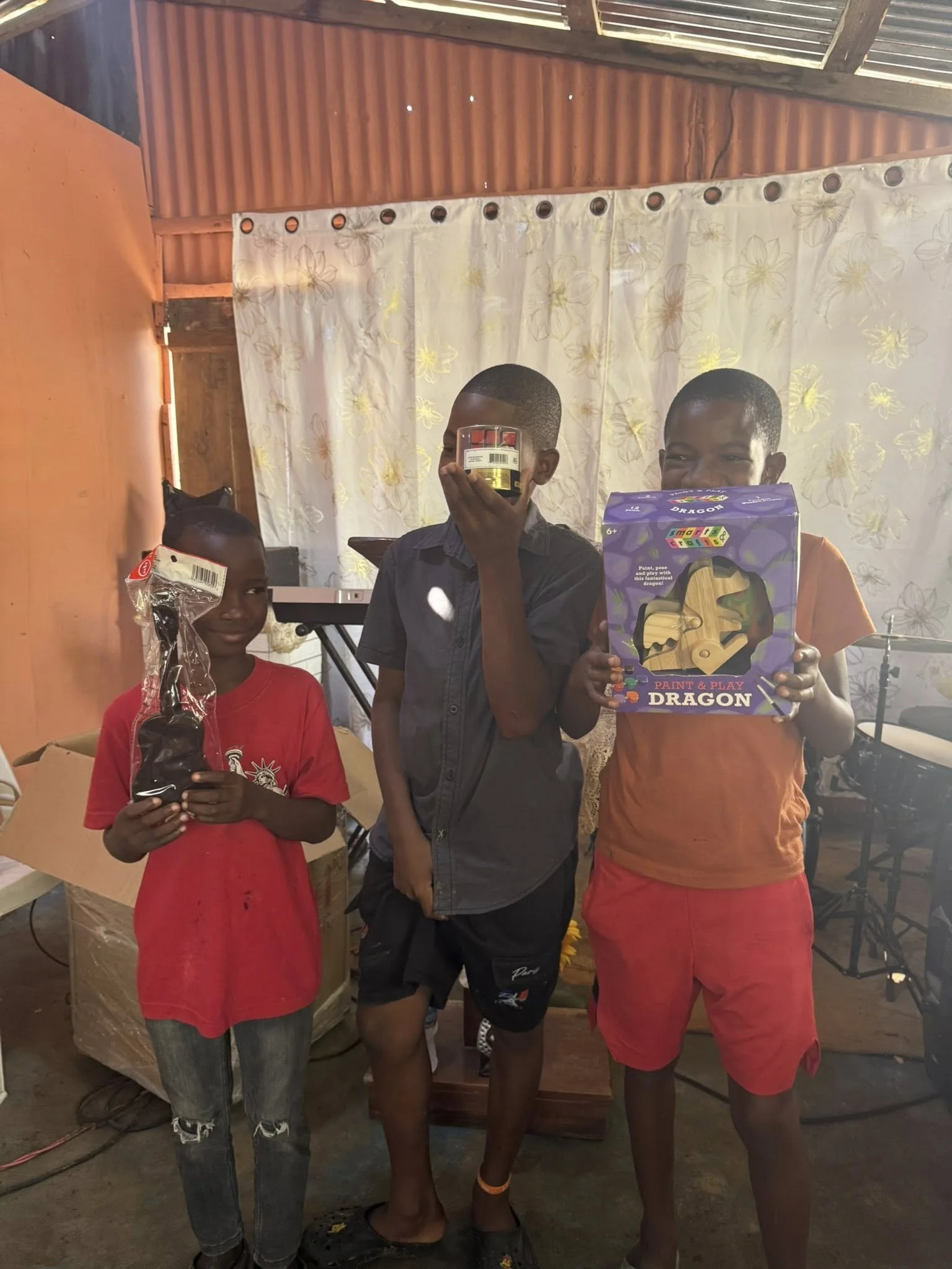 Three children standing indoors, each holding a different toy. The girl on the left is holding a wrapped toy; the boy in the middle is holding a small box covering his face; the boy on the right is holding a colorful box labeled 'Paint & Play Dragon'