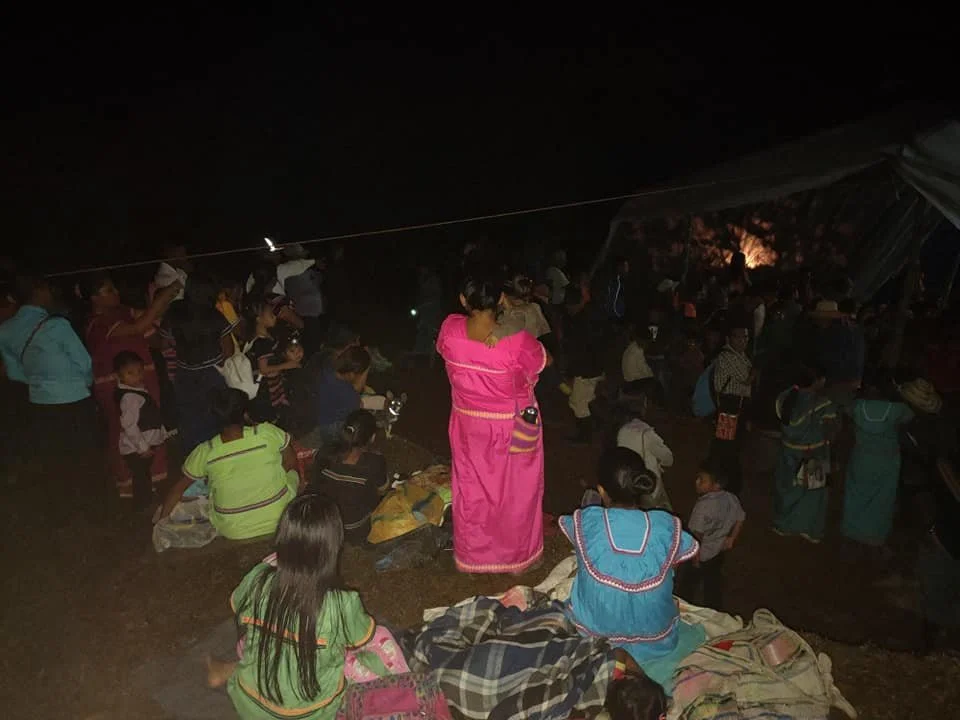 People gathered outdoors at night, some sitting and some standing, with a tent in the background.