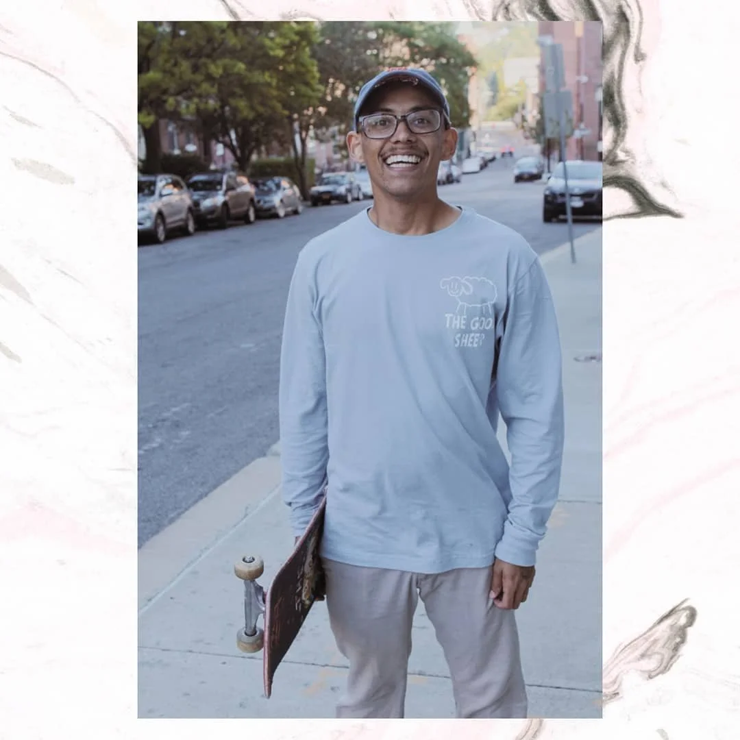 A man smiling while holding a skateboard on a city sidewalk with parked cars and trees in the background.