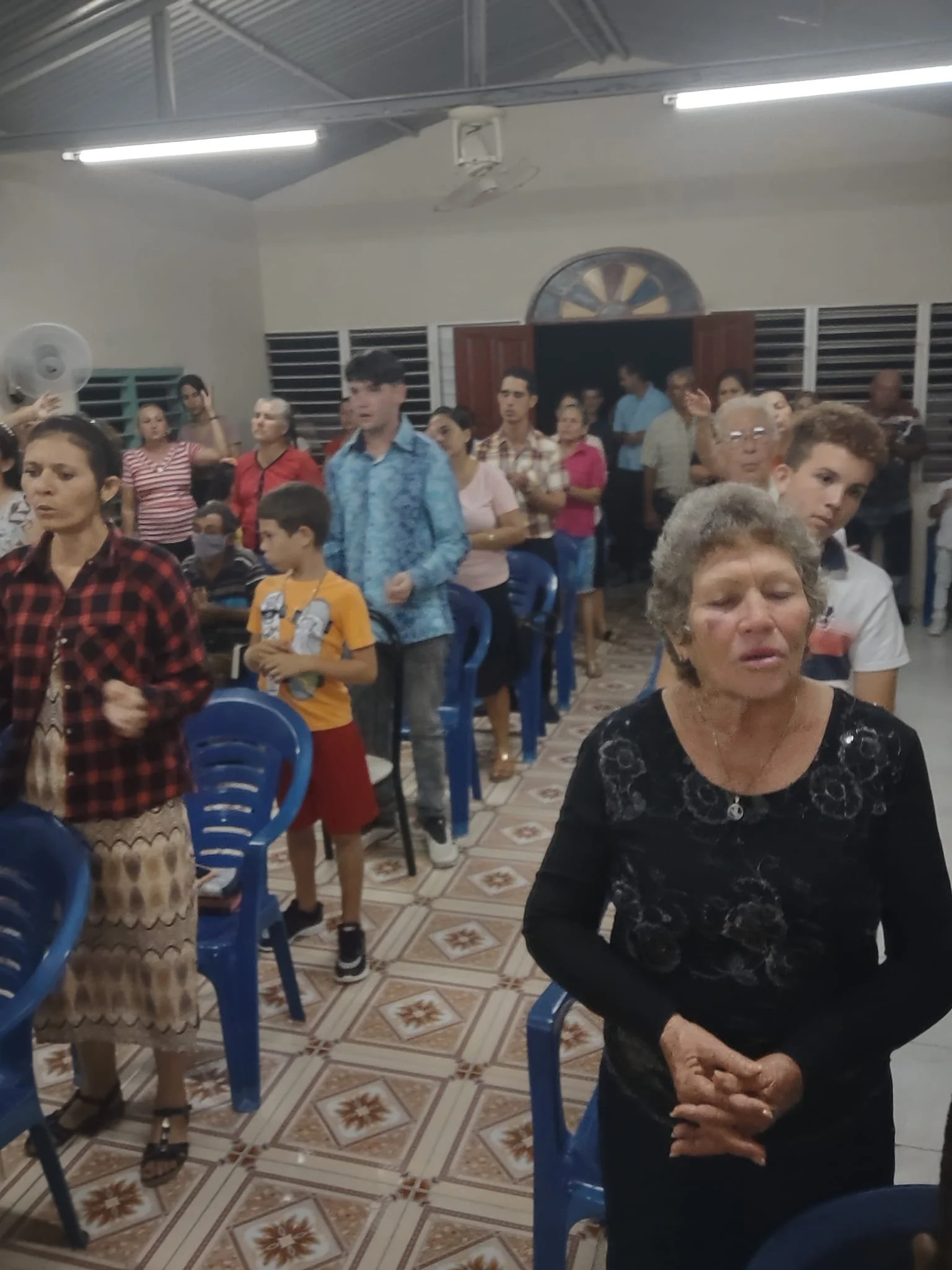 People gathered inside a room with blue plastic chairs, some standing, some sitting. The room has white walls, a tiled floor with a geometric pattern, and a high ceiling with fluorescent lights. Several individuals appear to be praying or reflecting,