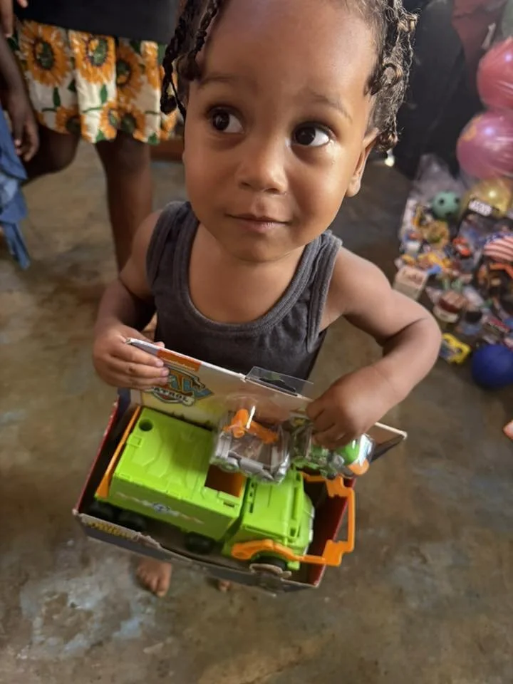 A young child with curly hair is holding a toy truck and standing on a concrete floor, with other children and balloons in the background.