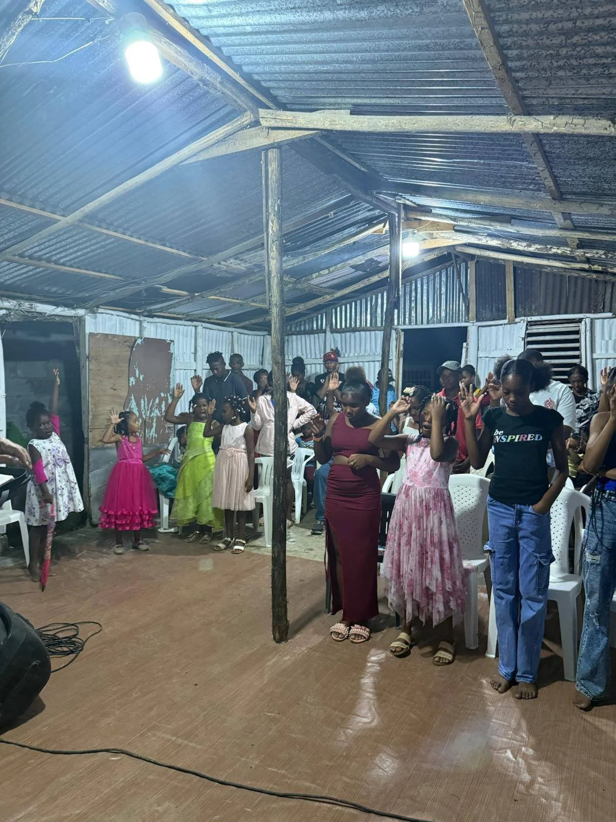 Group of children and adults standing indoors in a church or community hall, raising their hands, some with eyes closed, engaged in prayer or worship.