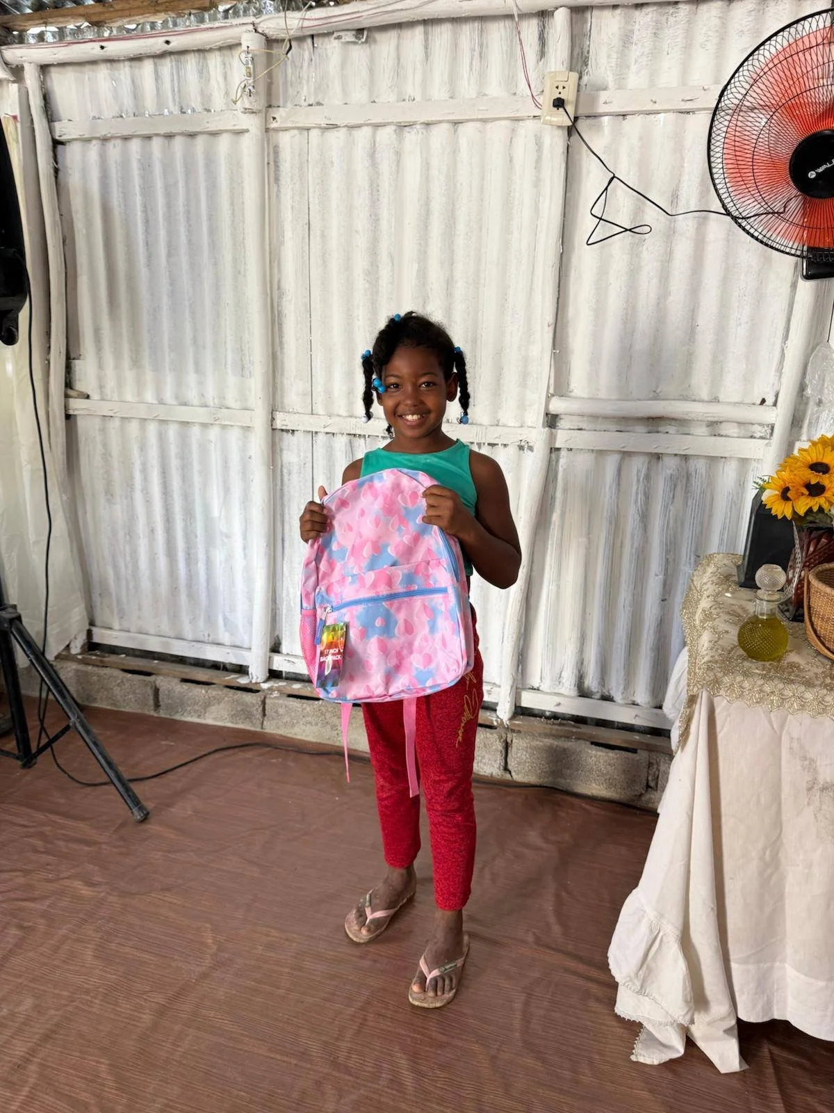 A smiling young girl with braided hair holding a pink backpack in a room with wooden flooring, a white painted corrugated metal wall, a standing fan, a table with a lace tablecloth, a vase with yellow flowers, and some electronic wires.