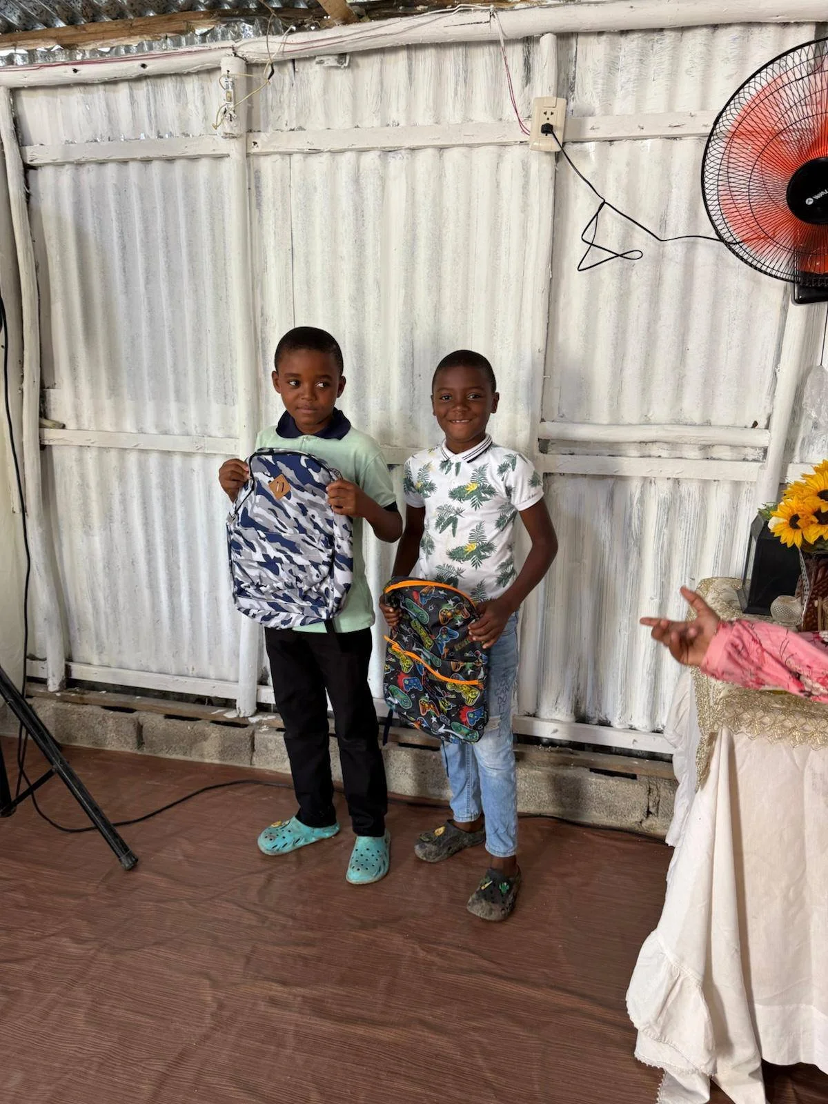 Two young boys standing indoors with corrugated metal wall and wooden floor, each holding a backpack, one wearing a light green shirt, the other in a white shirt with a tropical print, in a setting that appears to be a celebration or school event.