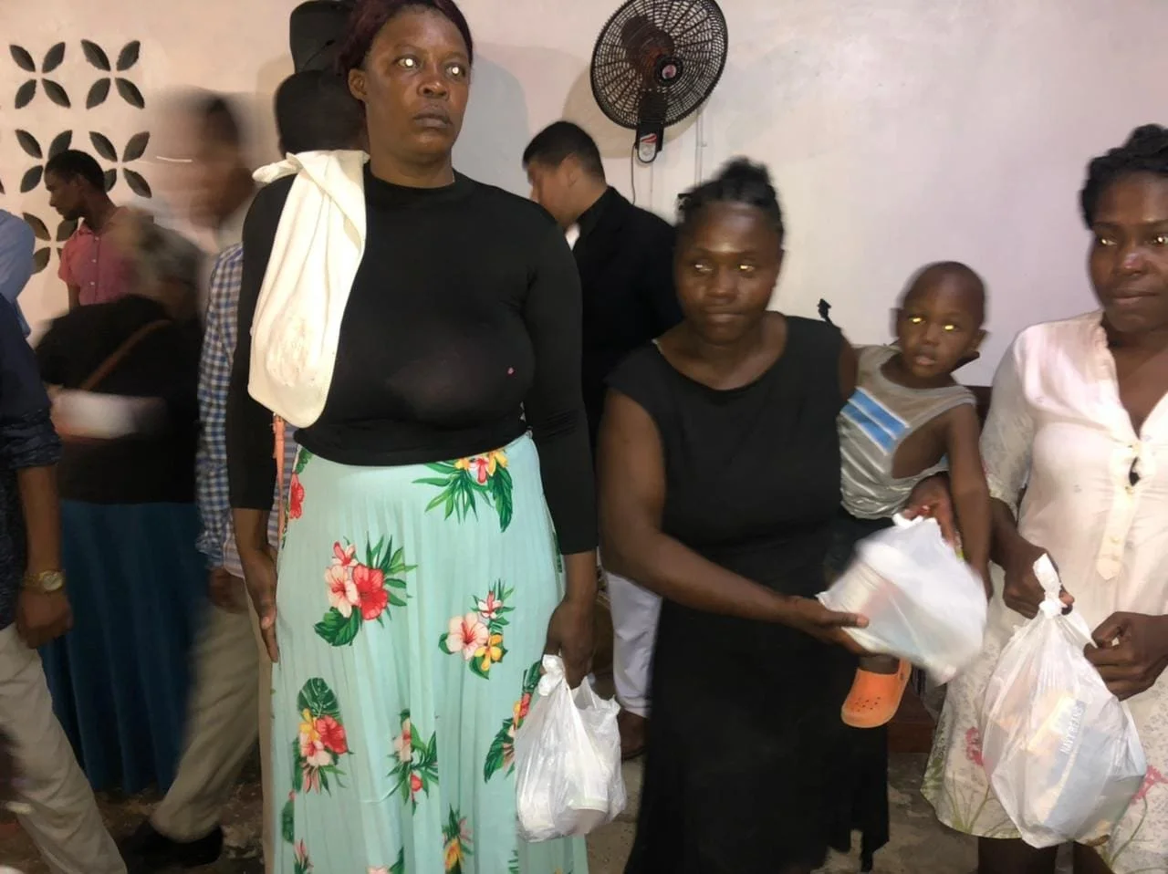 Group of people standing indoors, some holding plastic bags, with a wall-mounted fan in the background.